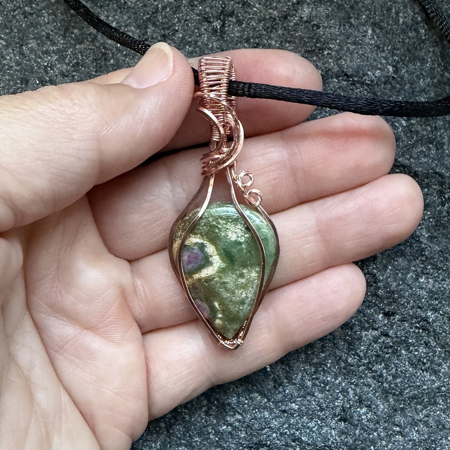 Front view of a handmade teardrop-shaped pendant necklace featuring a green ruby fuchsite stone wrapped in copper wire, suspended from a black cord, held in the artist's hand.