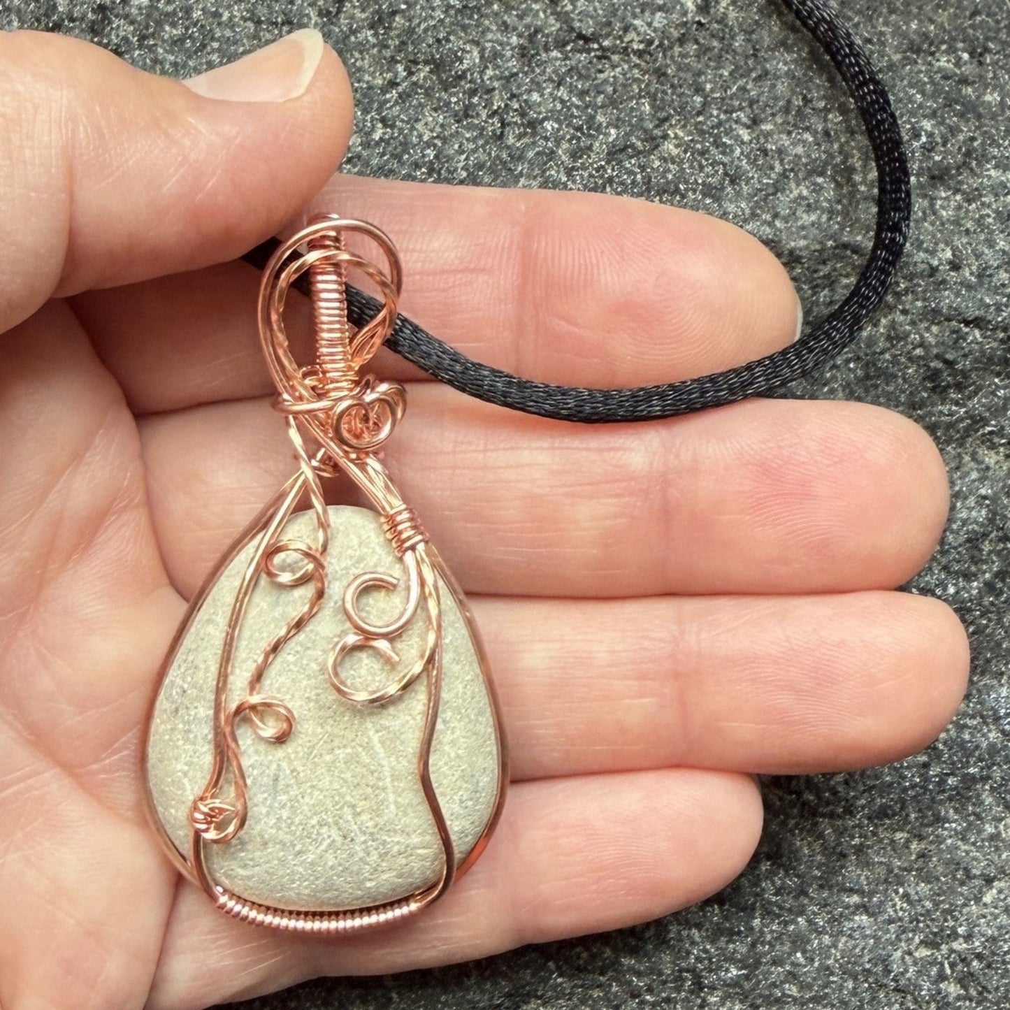 Front view of a teardrop-shaped stone pendant wrapped in copper wire, hanging from a black cord, held in the artist's hand.