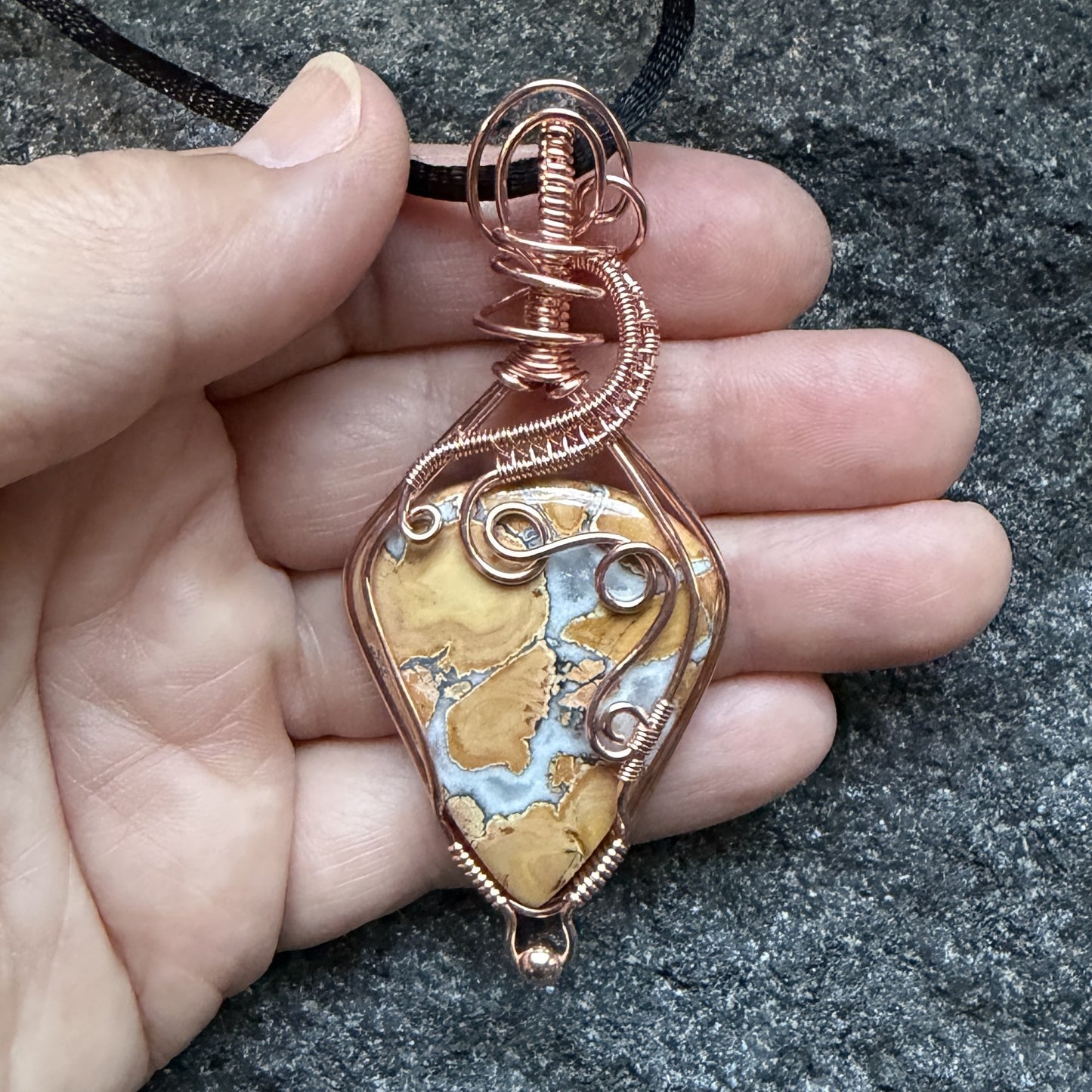 Front view of a handmade wire-wrapped pendant necklace featuring a polished yellow and white marbled maligano jasper stone, wrapped in copper wire with decorative swirls, hanging from a black cord, held in the artist's hand.