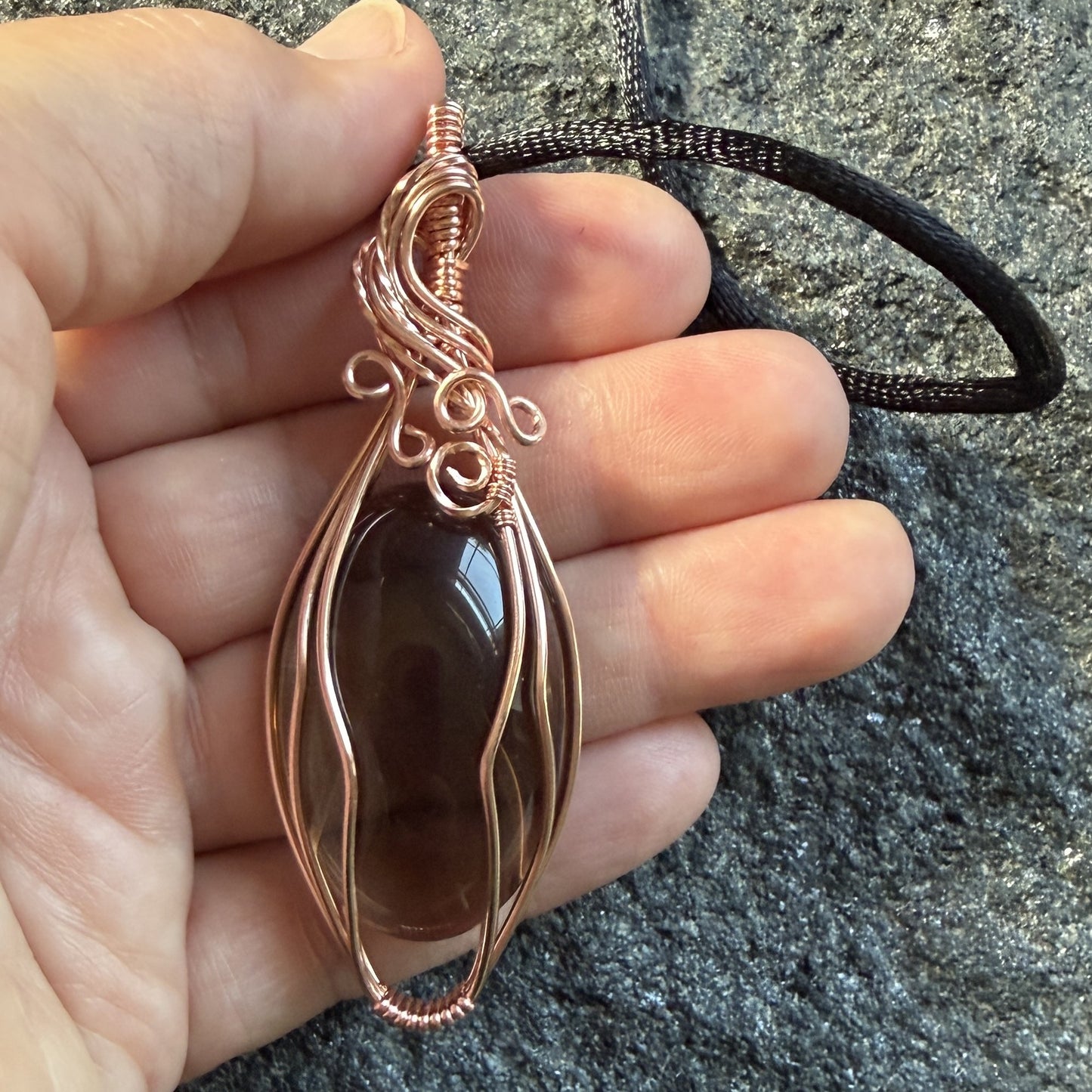Front view of a teardrop-shaped dark brown smoky quartz stone pendant wrapped in copper wire, hanging from a black cord, held in the artist's hand.