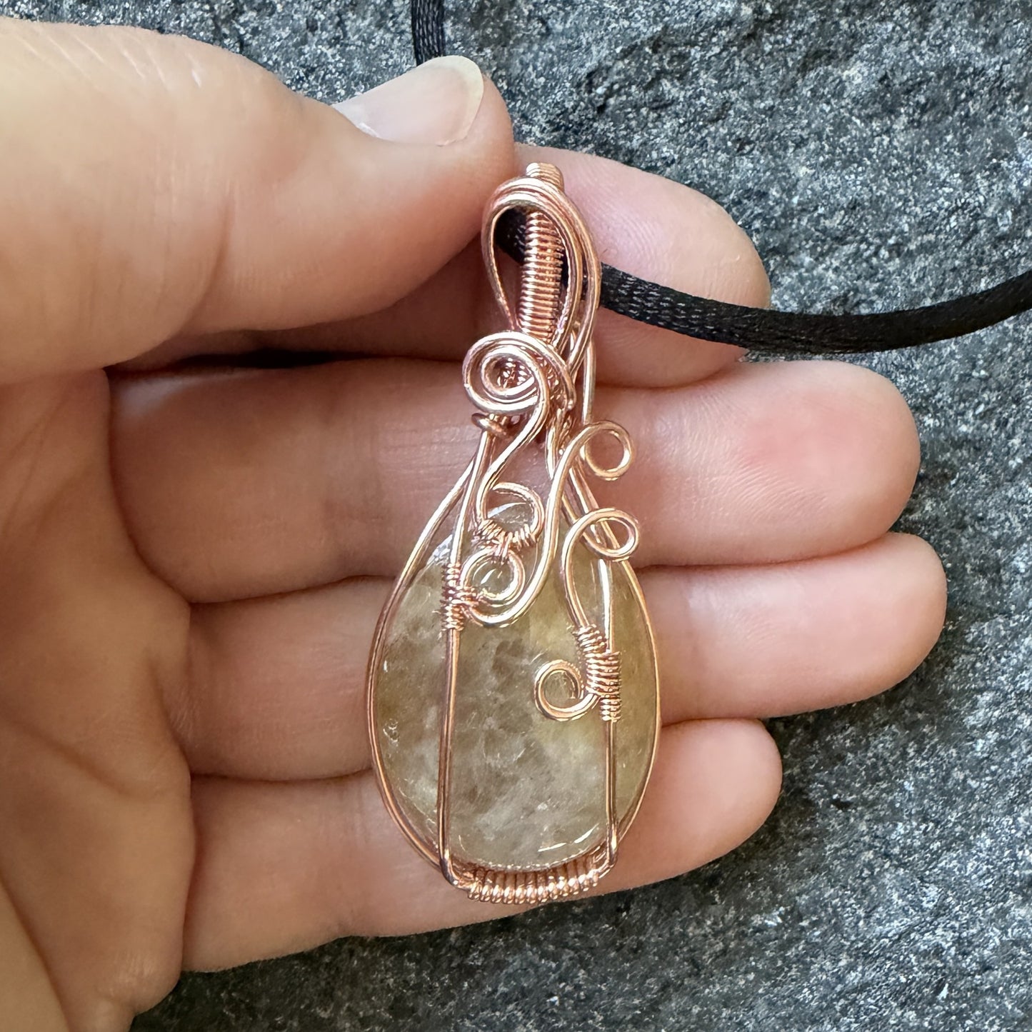 Front view of a teardrop-shaped pendant necklace featuring a pale yellow quartz stone wrapped in copper wire, held in the artist's hand.