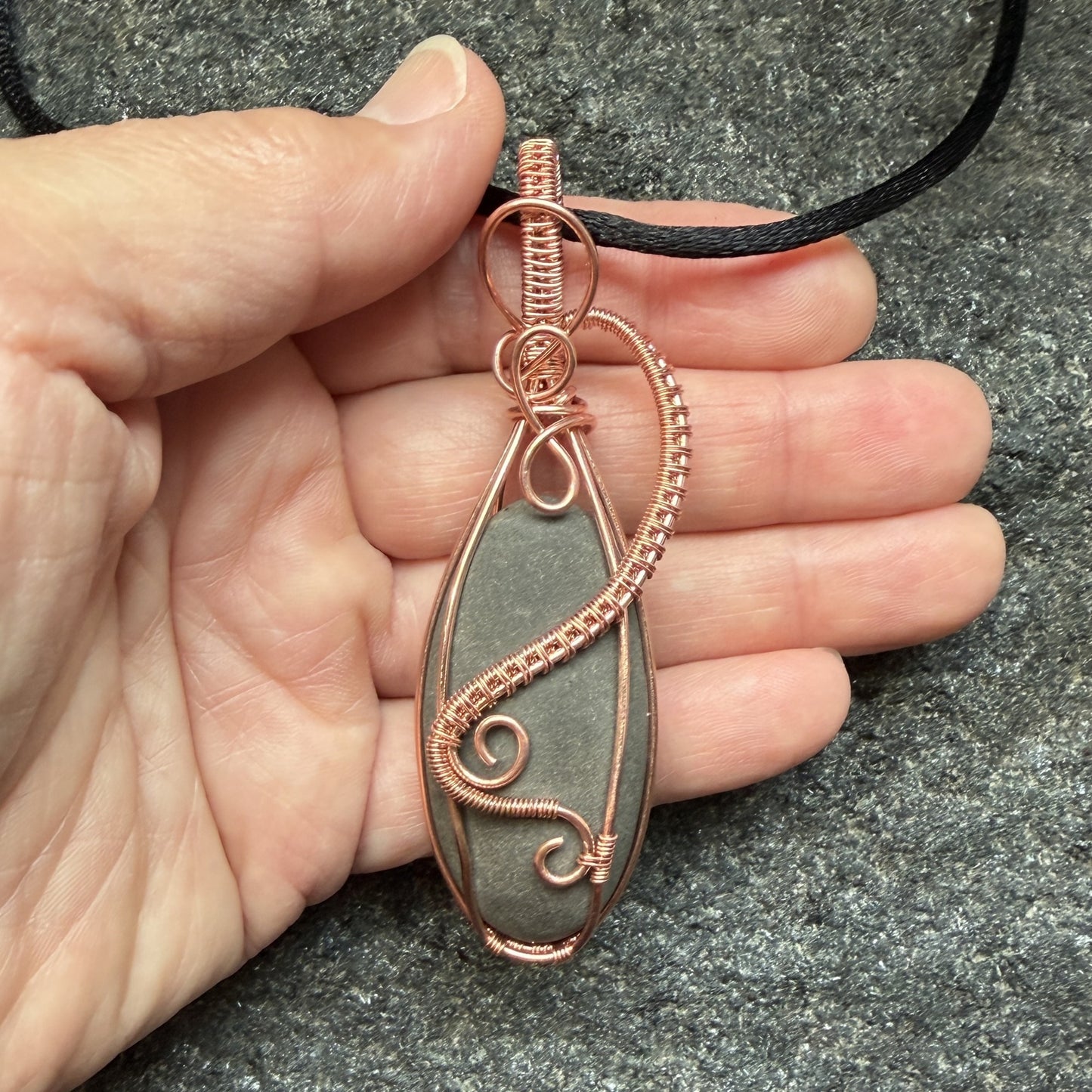 Front view of a narrow teardrop-shaped pendant necklace featuring a dark gray stone wrapped in copper wire, suspended from a black cord, held in the artist's hand.