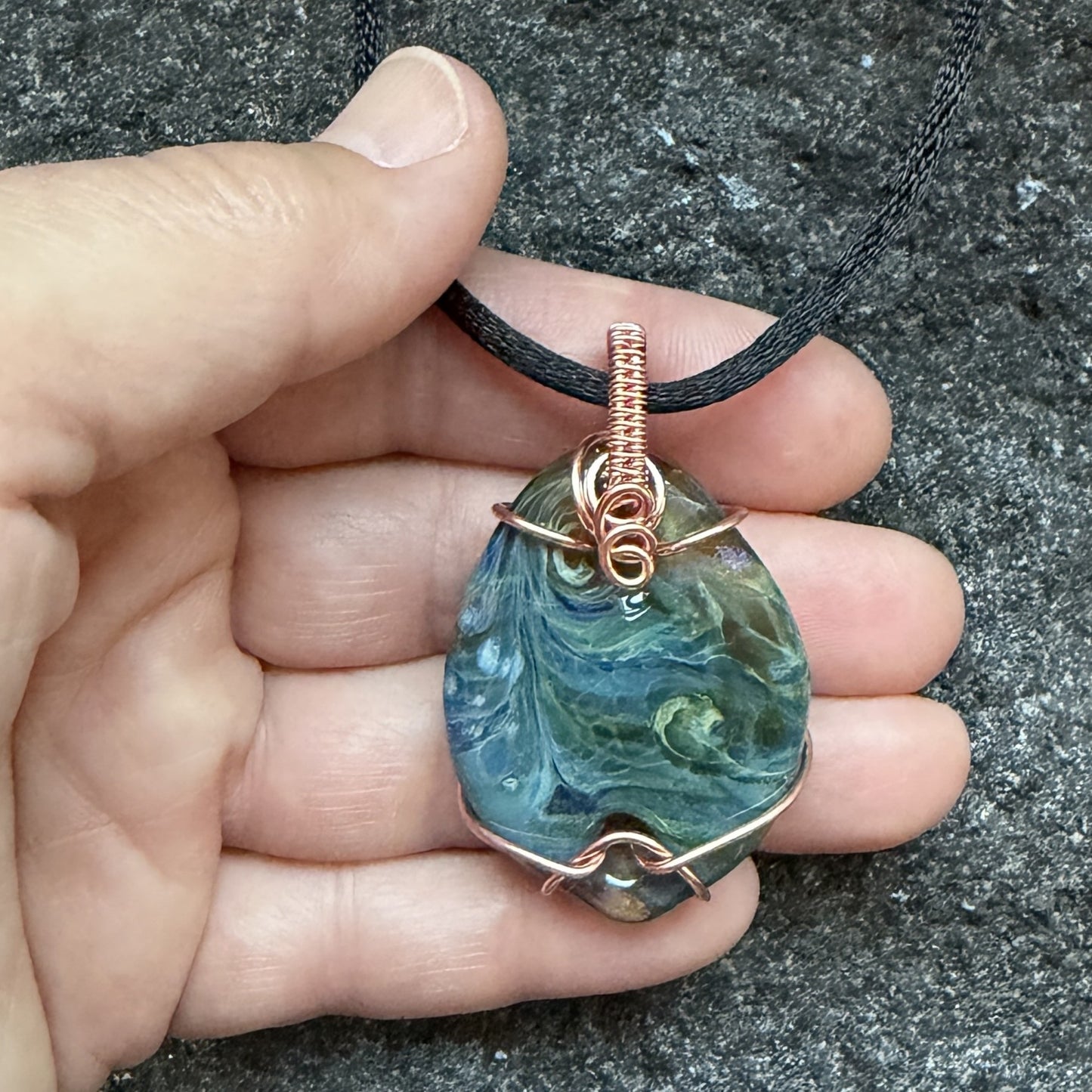 Front view of side 2 of a handmade pendant necklace featuring a marbled blue and green glass stone wrapped in copper wire, hanging from a black cord held in the artist's hand.