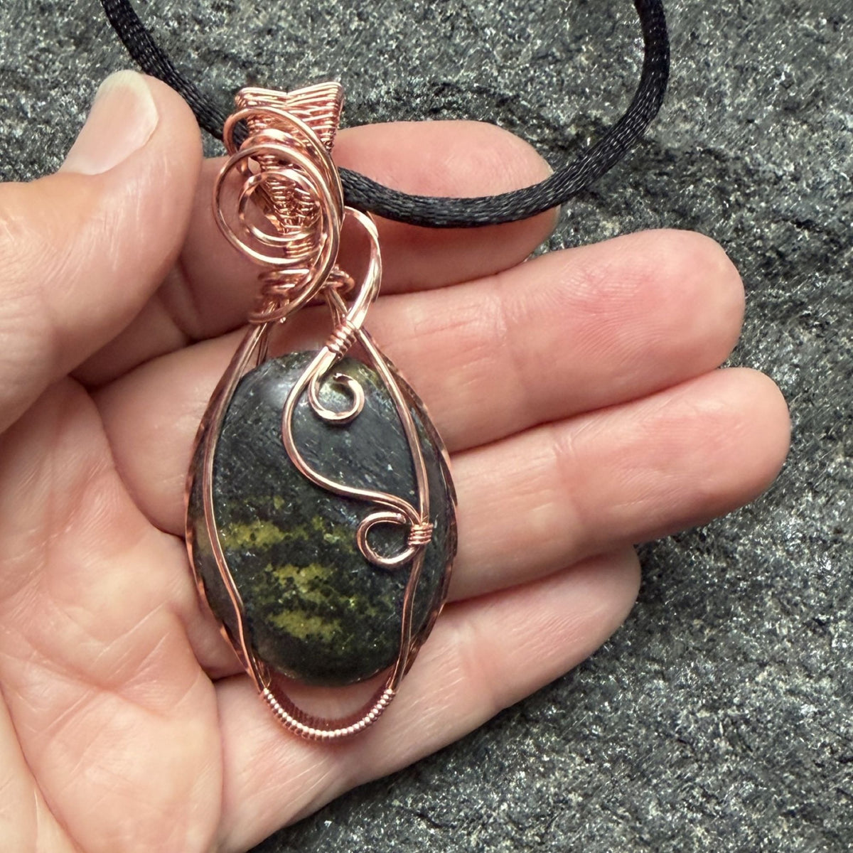 Front view of a teardrop-shaped wire-wrapped pendant necklace featuring a dark stone with olive green inclusions, set in copper wire and suspended from a black cord, shown held in the artist's hand.