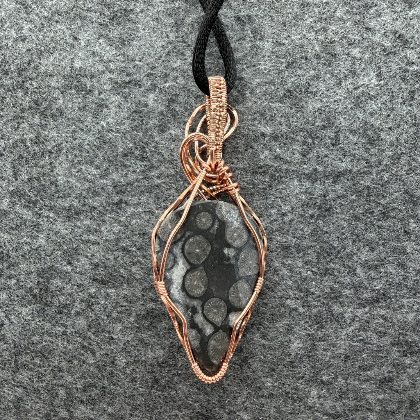Back view of a handmade pendant featuring a black coral fossil stone wrapped in copper wire, hanging from a black cord against a textured gray felt background.