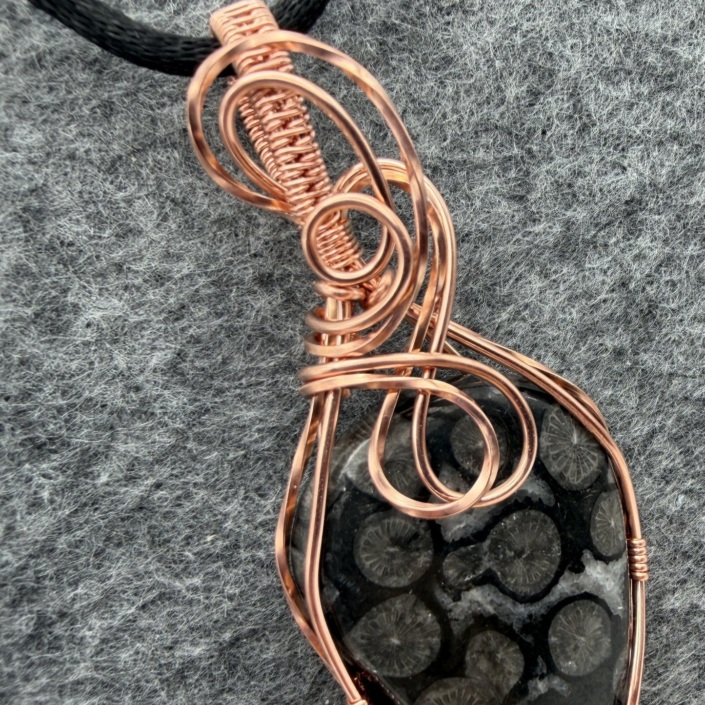 Detail view of a handmade pendant featuring a black coral fossil stone wrapped in copper wire, hanging from a black cord against a textured gray felt background.