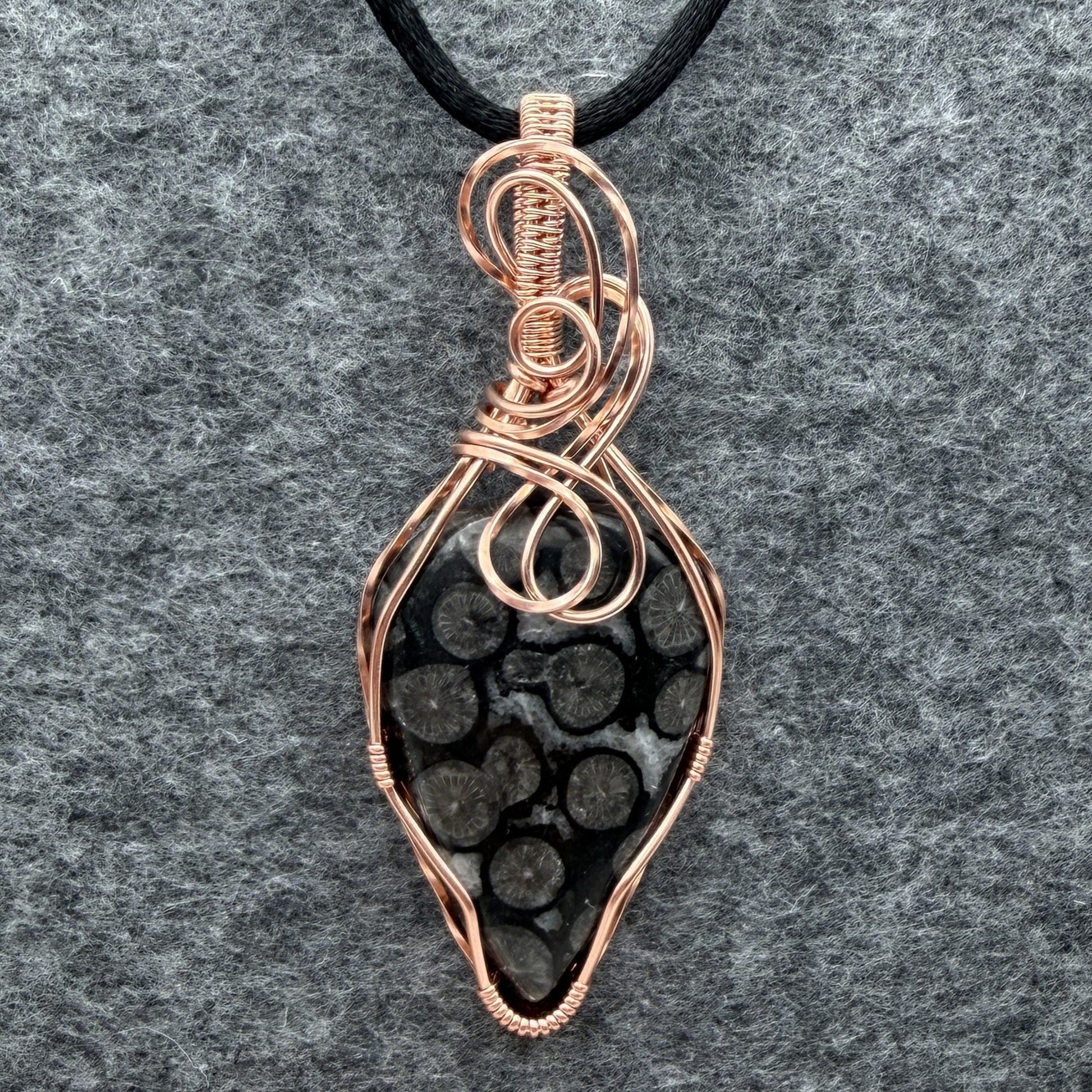Front view of a handmade pendant featuring a black coral fossil stone wrapped in copper wire, hanging from a black cord against a textured gray felt background.