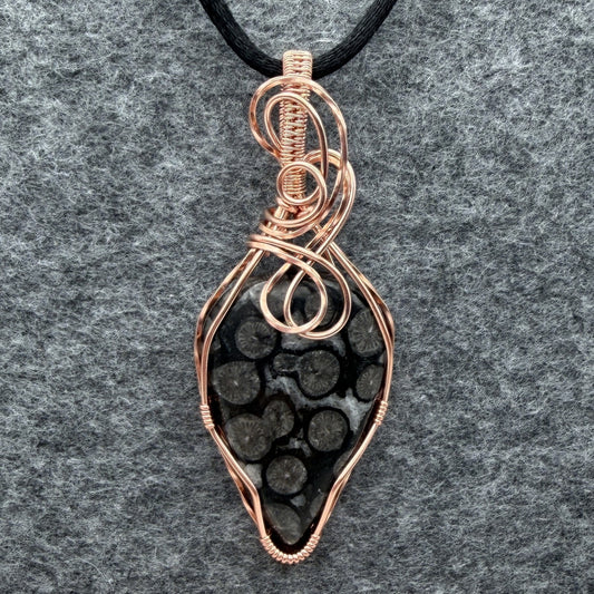 Front view of a handmade pendant featuring a black coral fossil stone wrapped in copper wire, hanging from a black cord against a textured gray felt background.