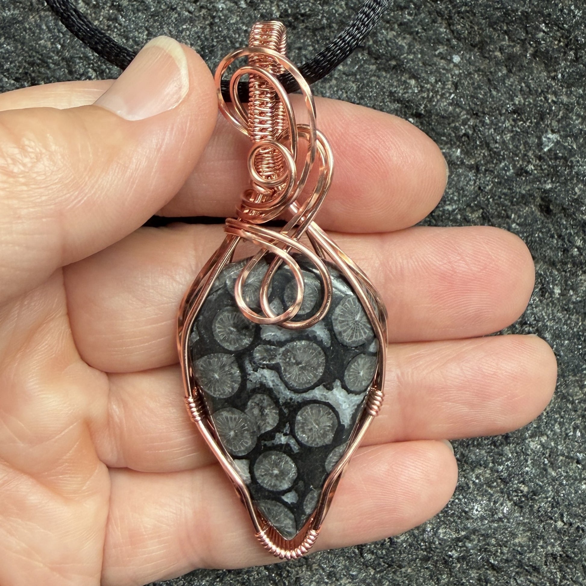 Front view of a handmade pendant featuring a black coral fossil stone wrapped in copper wire, hanging from a black cord, held in the artist's hand.