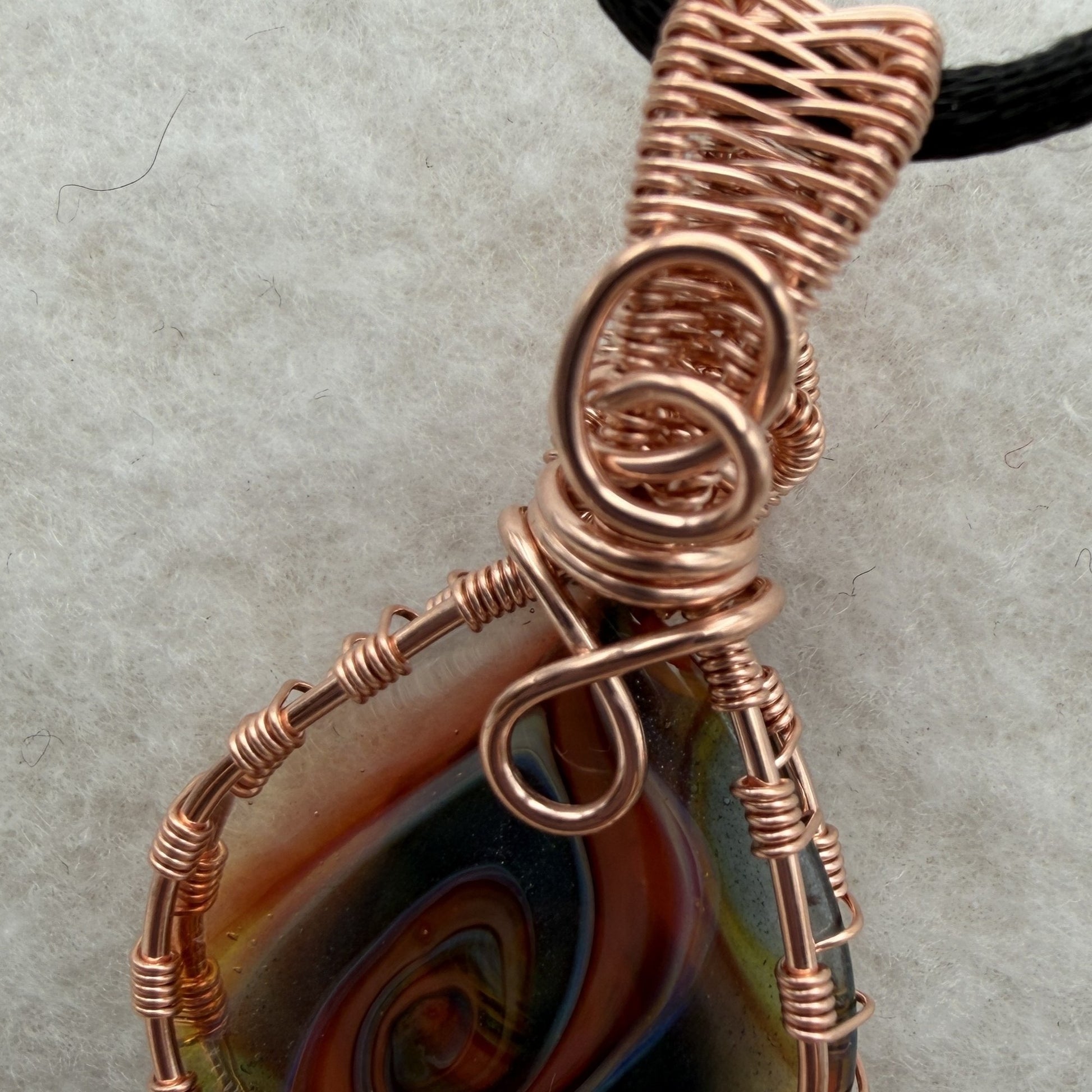 Detail view of a reversible teardrop-shaped pendant necklace featuring a marbled swirl of brown, orange, green, and blue glass, wrapped in copper-colored wire and suspended from a black cord, set against a white felt background.