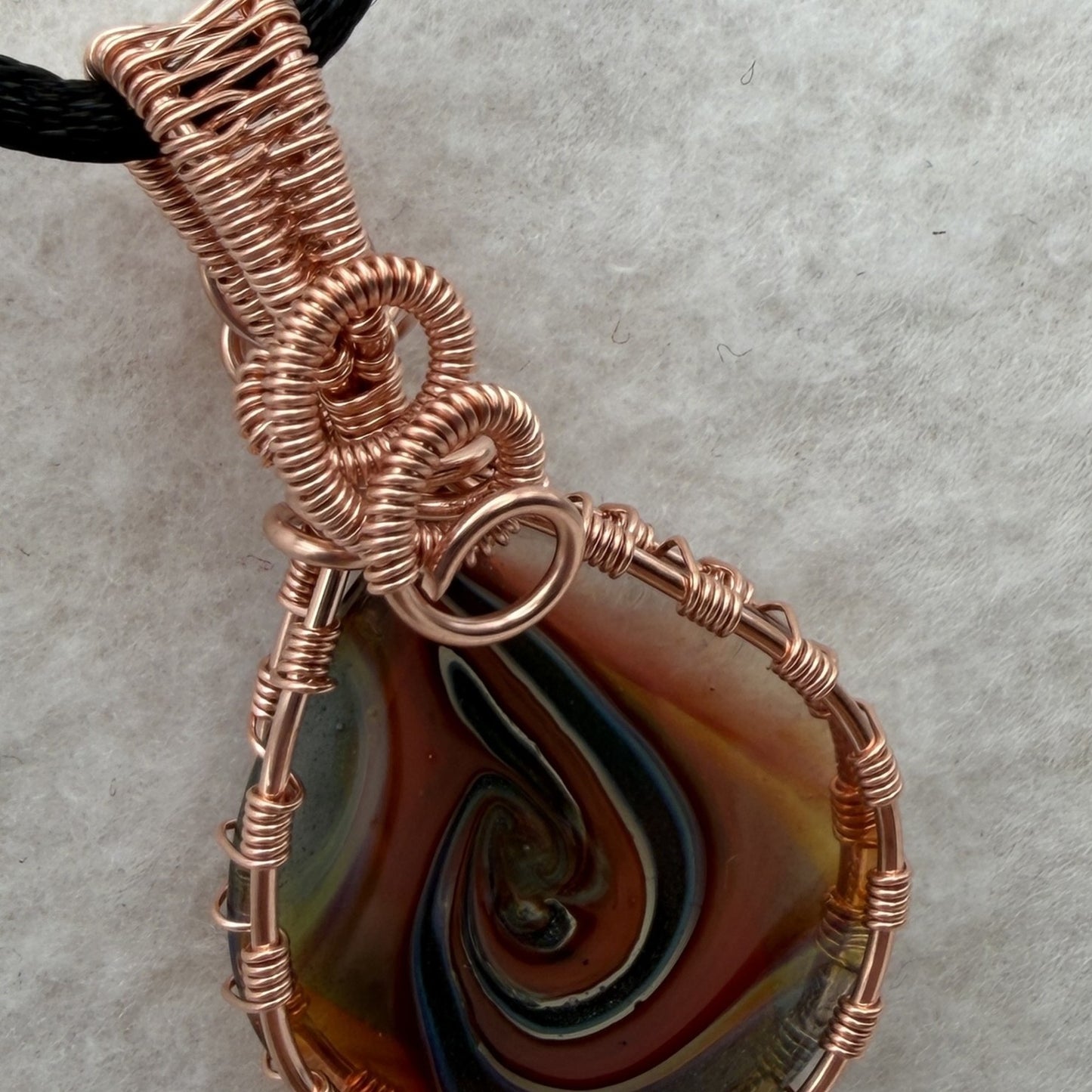 Detail view of a reversible teardrop-shaped pendant necklace featuring a marbled swirl of brown, orange, green, and blue glass, wrapped in copper-colored wire and suspended from a black cord, set against a cream background.