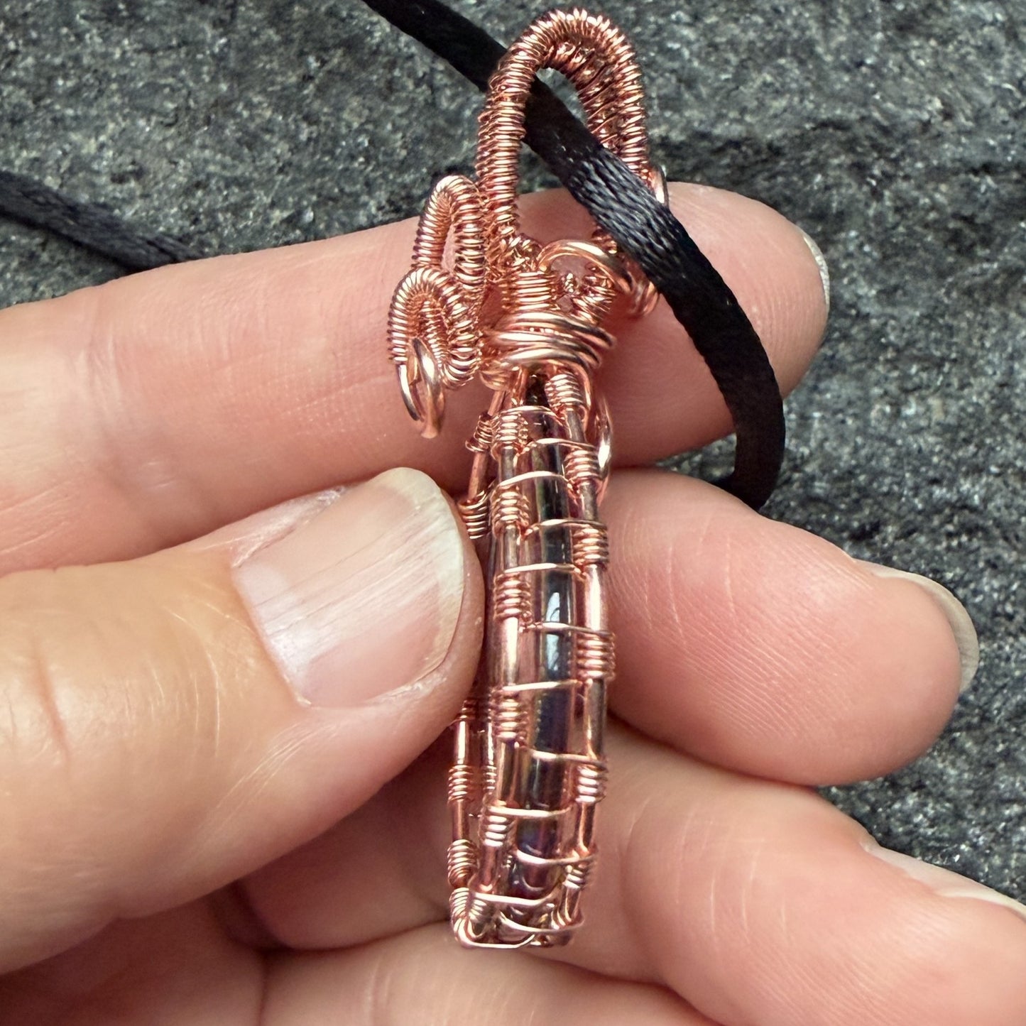 Detail view of a reversible teardrop-shaped pendant necklace featuring a marbled swirl of brown, orange, green, and blue glass, wrapped in copper-colored wire and suspended from a black cord, set against a cream background.