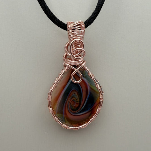 Front view of a reversible teardrop-shaped pendant necklace featuring a marbled swirl of brown, orange, green, and blue glass, wrapped in copper-colored wire and suspended from a black cord, set against a cream background.
