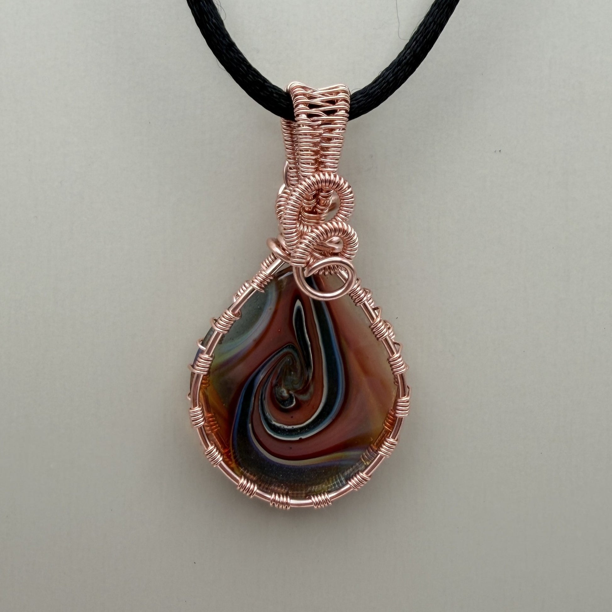 Back view of a reversible teardrop-shaped pendant necklace featuring a marbled swirl of brown, orange, green, and blue glass, wrapped in copper-colored wire and suspended from a black cord, set against a cream background.