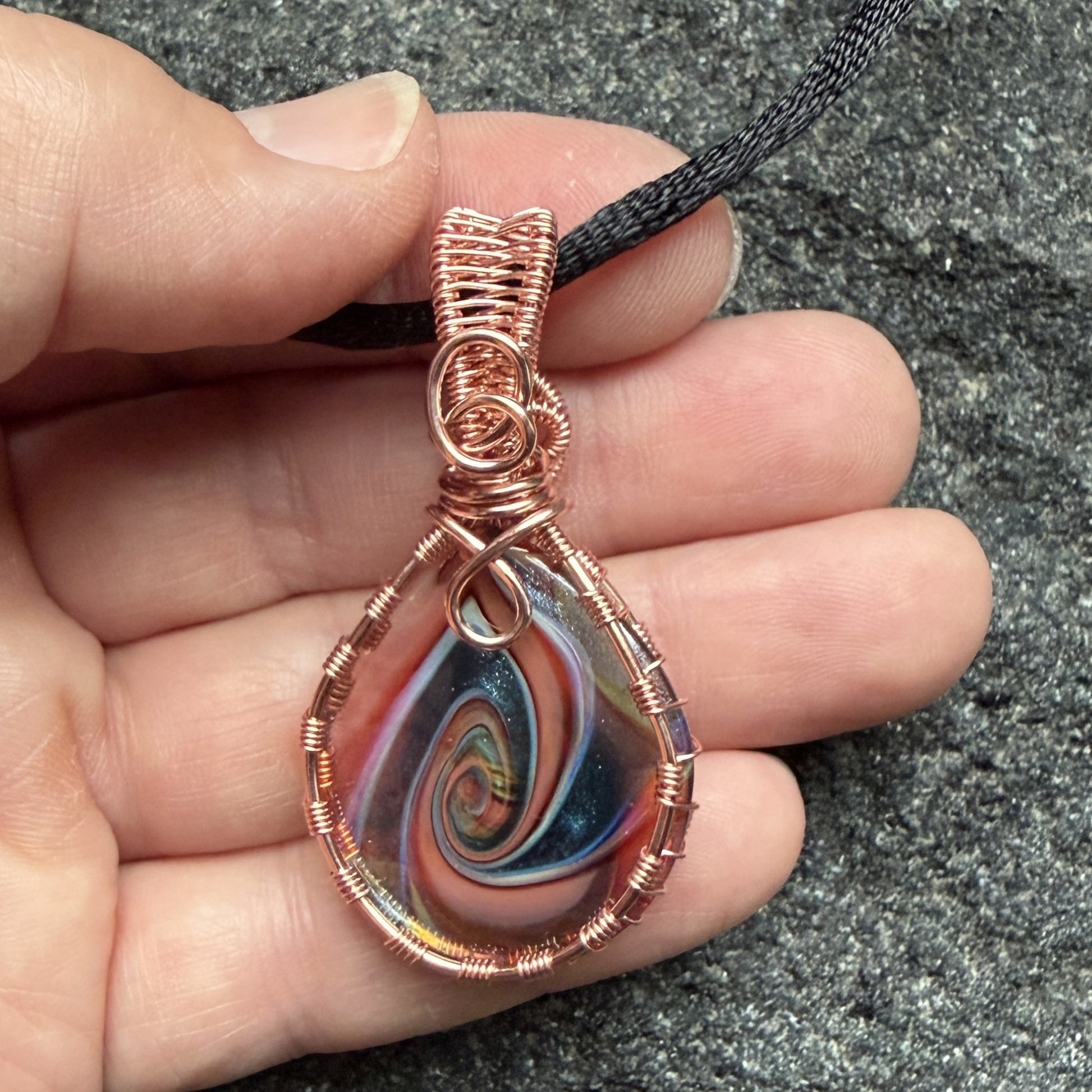Front view of a reversible teardrop-shaped pendant necklace featuring a marbled swirl of brown, orange, green, and blue glass, wrapped in copper-colored wire and suspended from a black cord, held in the artist's hand.