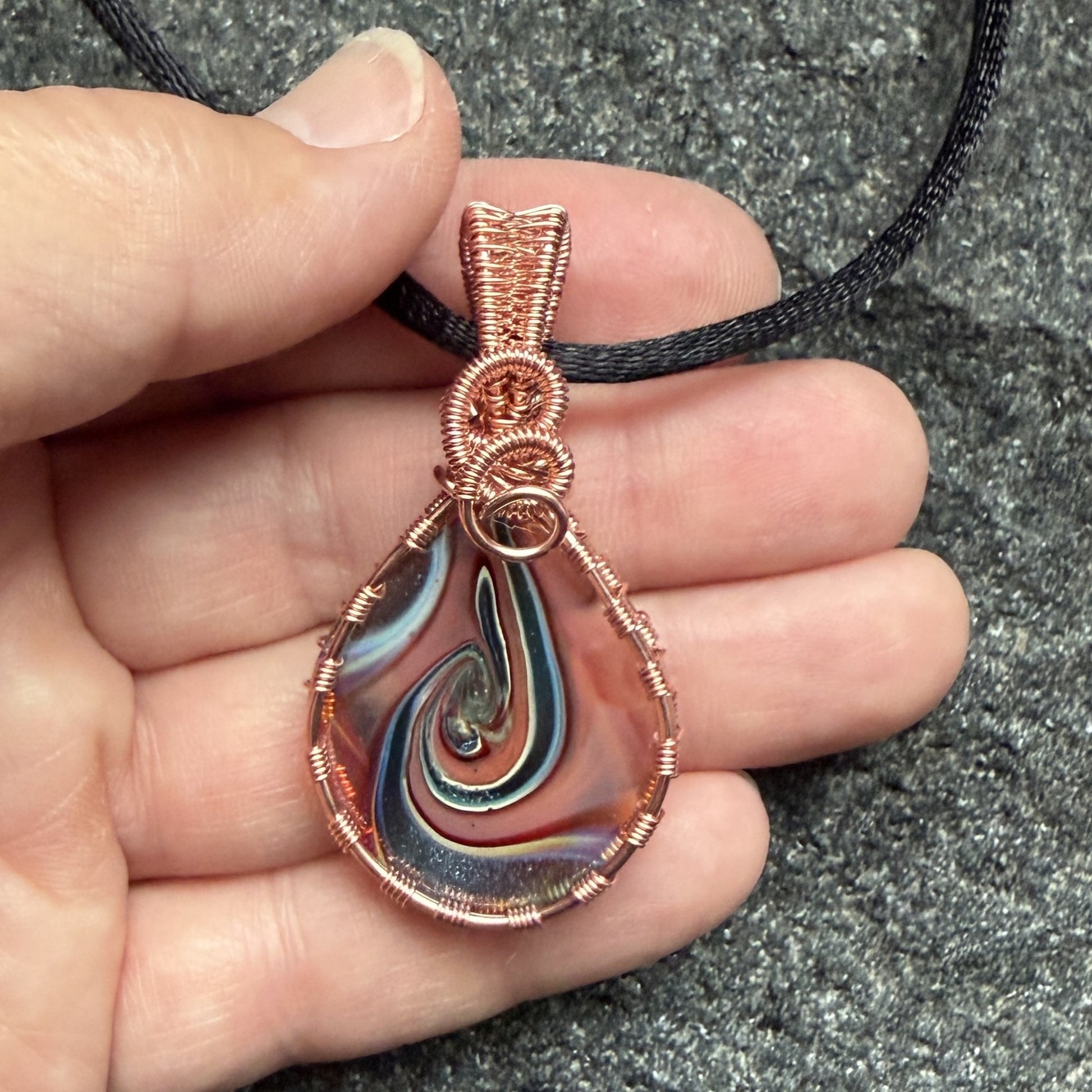 Back view of a reversible teardrop-shaped pendant necklace featuring a marbled swirl of brown, orange, green, and blue glass, wrapped in copper-colored wire and suspended from a black cord, held in the artist's hand.