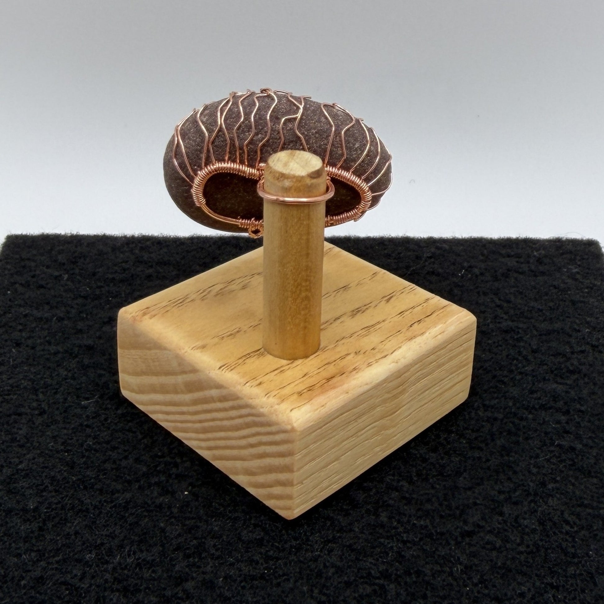 Back view of a decorative stone sculpture featuring a dark brown textured oval stone with a copper wire tree design, mounted on a light-colored wooden block against a split gray and black background.
