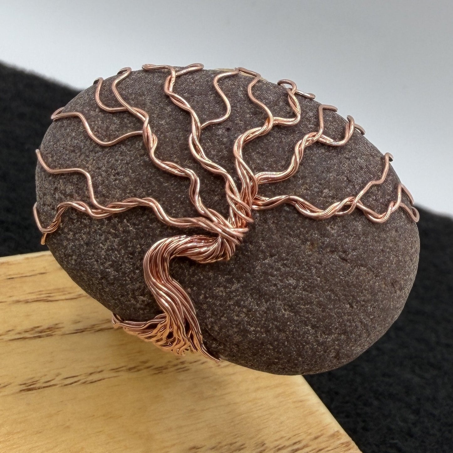 Detail view of a decorative stone sculpture featuring a dark brown textured oval stone with a copper wire tree design, mounted on a light-colored wooden block against a split gray and black background.
