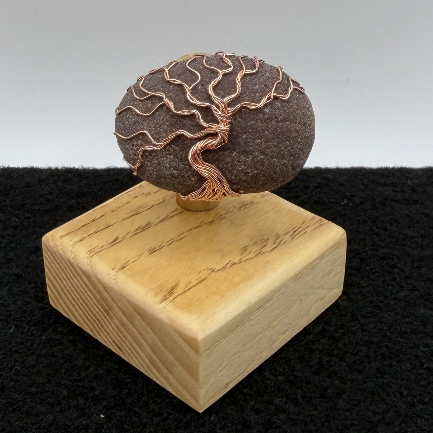 Front view of a decorative stone sculpture featuring a dark brown textured oval stone with a copper wire tree design, mounted on a light-colored wooden block against a split gray and black background.