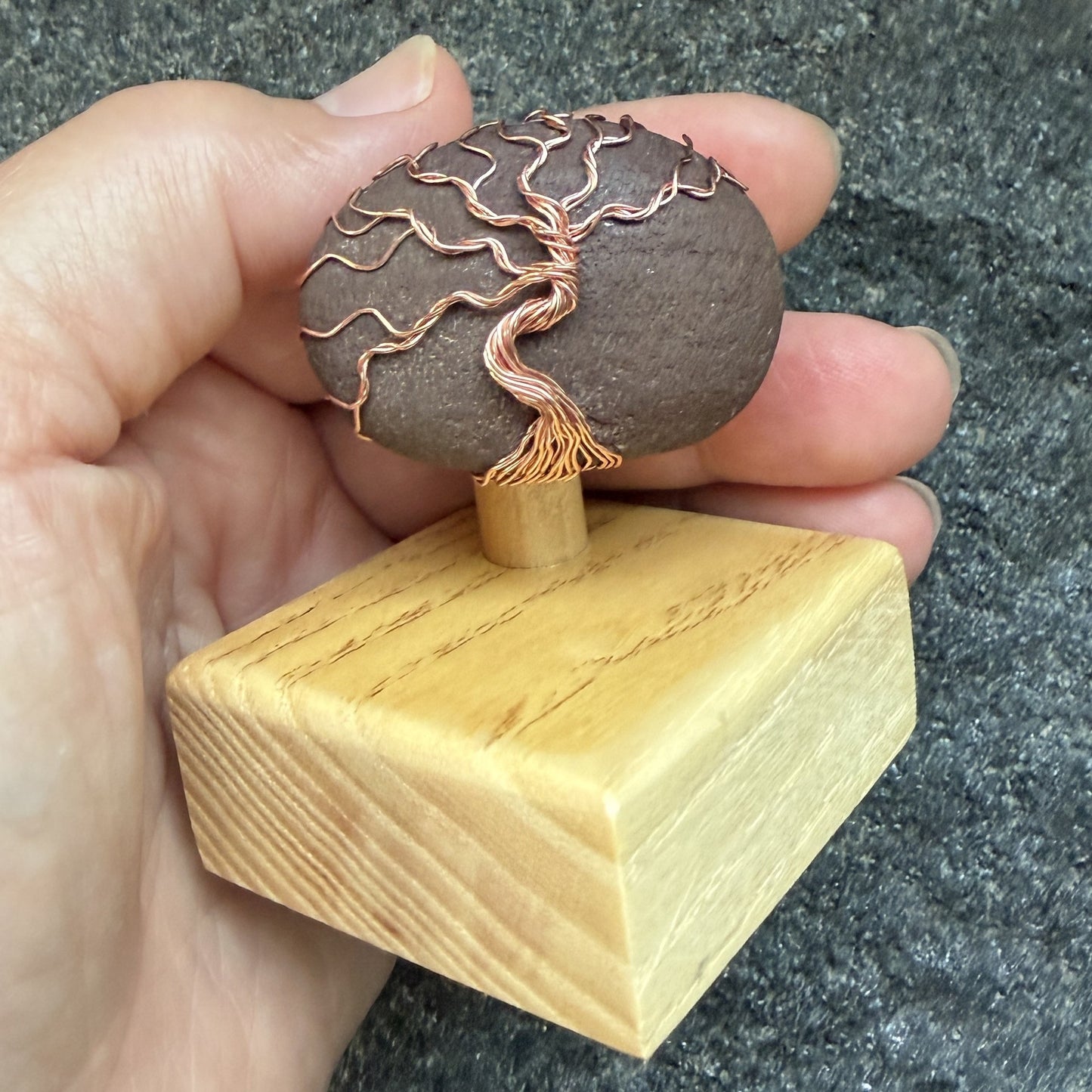 Front view of a decorative stone sculpture featuring a dark brown textured oval stone with a copper wire tree design, mounted on a light-colored wooden block held in the artist's hand.