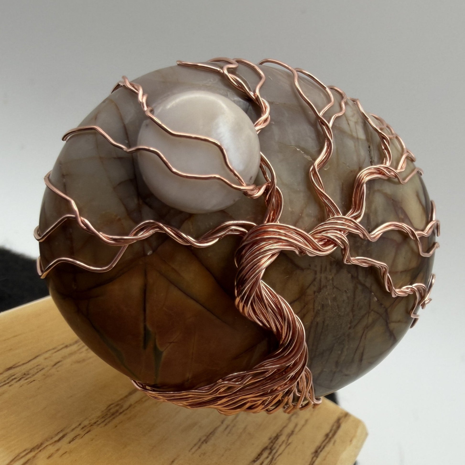 Detail view of a decorative tree-of-life sculpture made from polished picasso jasper and copper wire, mounted on a light-colored wooden block against a neutral gray background.