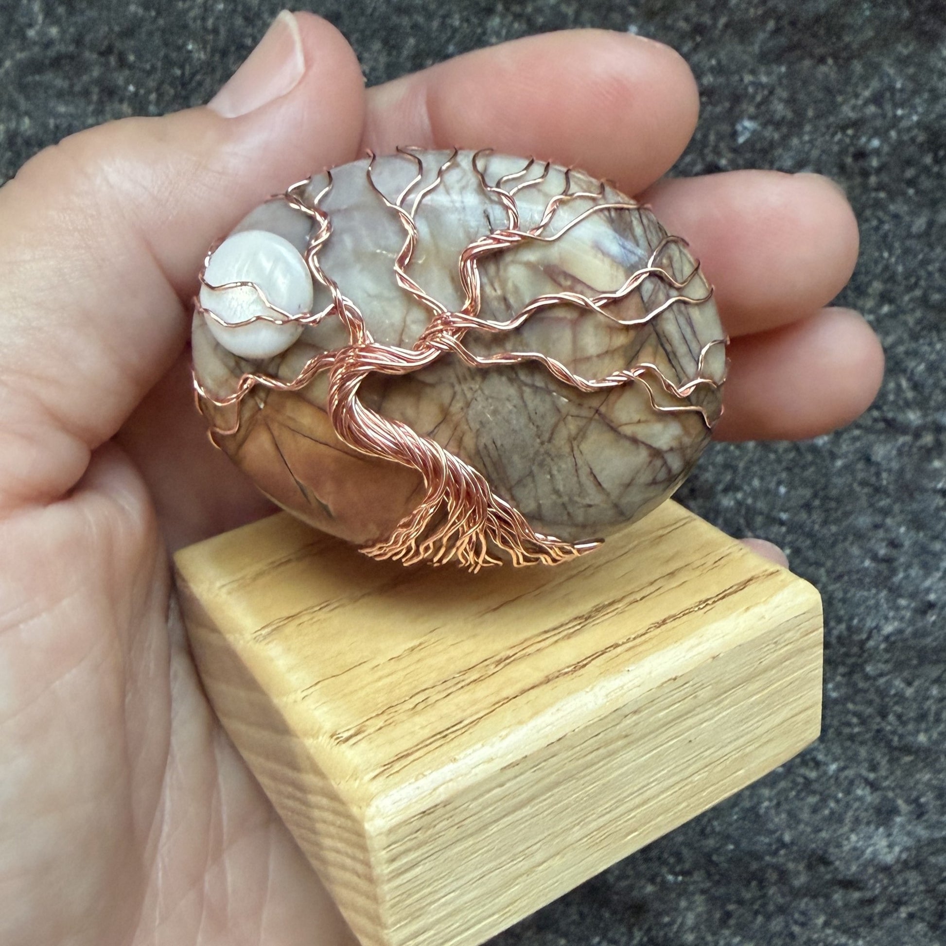 Front view of a decorative tree-of-life sculpture made from polished picasso jasper and copper wire, mounted on a light-colored wooden block held in the artist's hand.