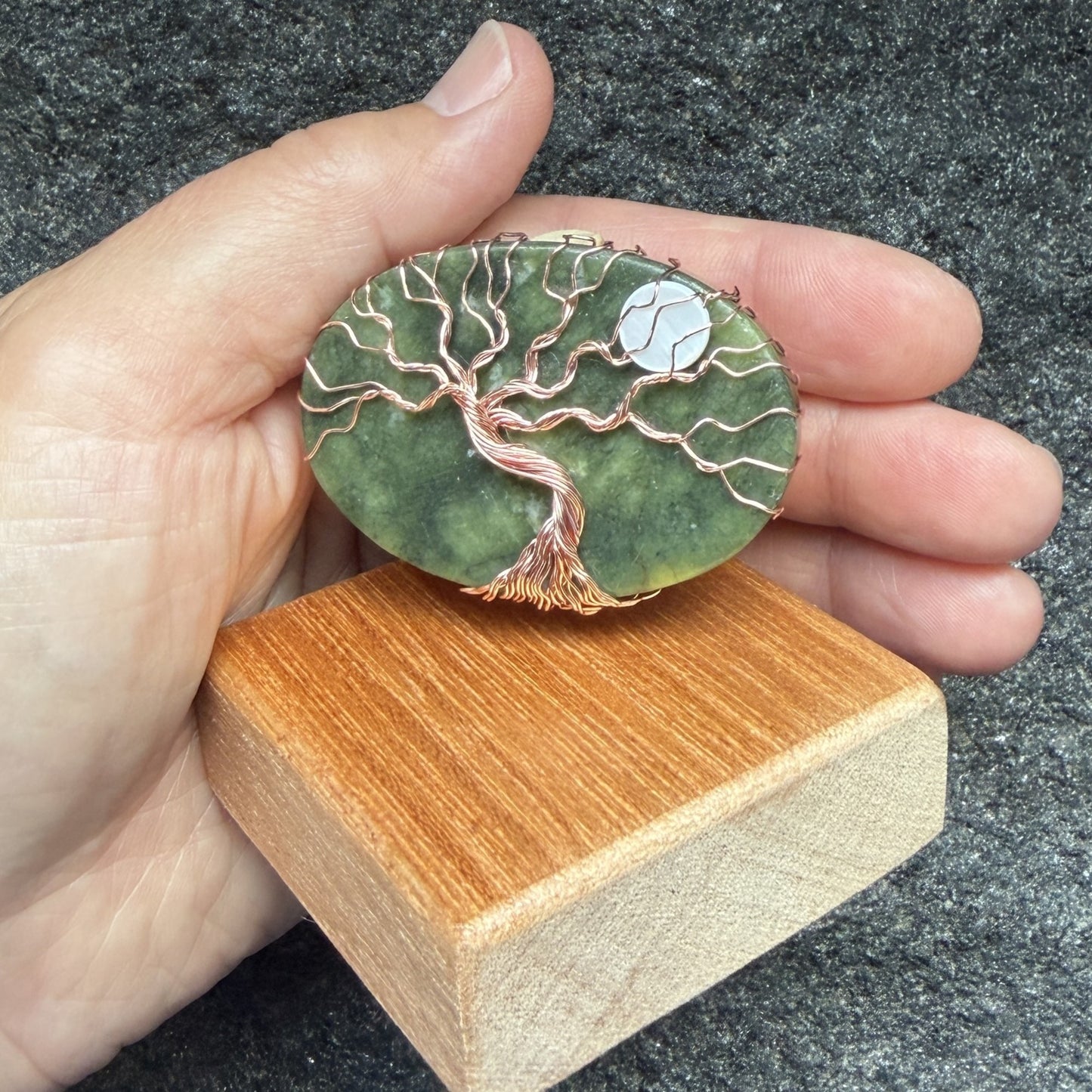 Front view of a tree of life display piece featuring a green serpentine stone with copper wire roots and branches and a white moon, mounted on a square wooden display block. Held in the artist's hand.
