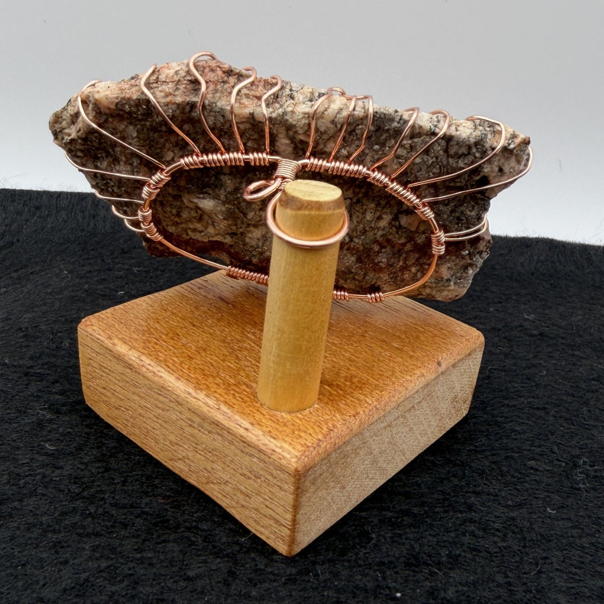 Back view of a decorative tree of life sculpture made from copper wire wrapped over a rough granite slab, displayed on a rectangular light wood block against a neutral gray and black background.