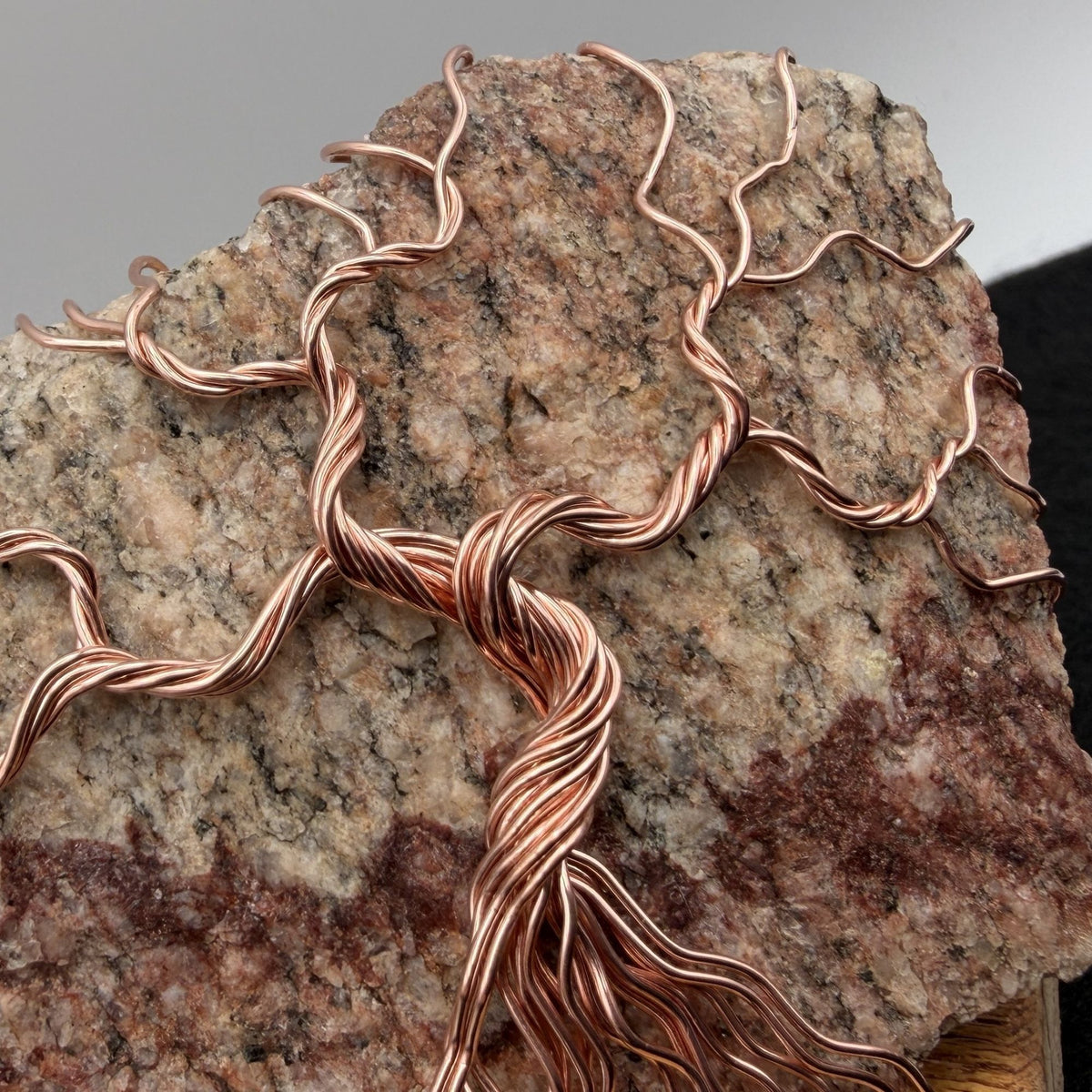 Detail view of a decorative tree of life sculpture made from copper wire wrapped over a rough granite slab, displayed on a rectangular light wood block against a neutral gray and black background.