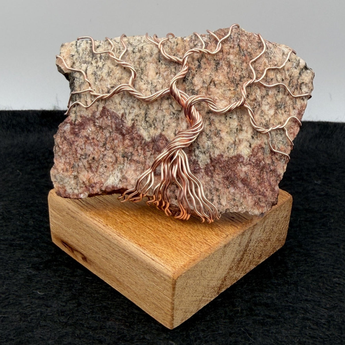 Front view of a decorative tree of life sculpture made from copper wire wrapped over a rough granite slab, displayed on a rectangular light wood block against a neutral gray and black background.