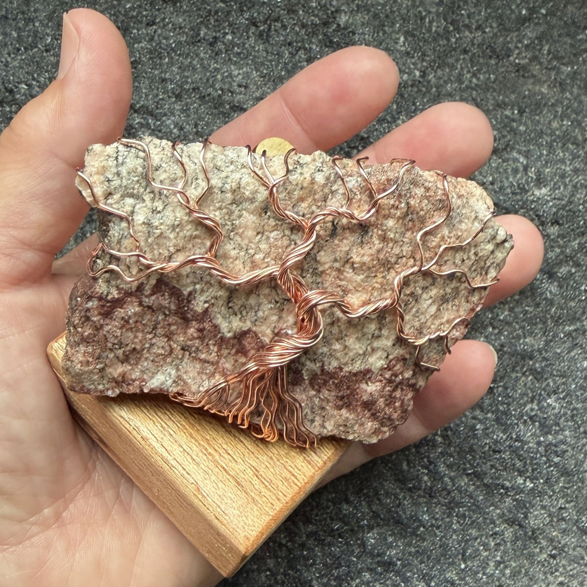 Front view of a decorative tree of life sculpture made from copper wire wrapped over a rough granite slab, displayed on a rectangular light wood block held in the artist's hand.