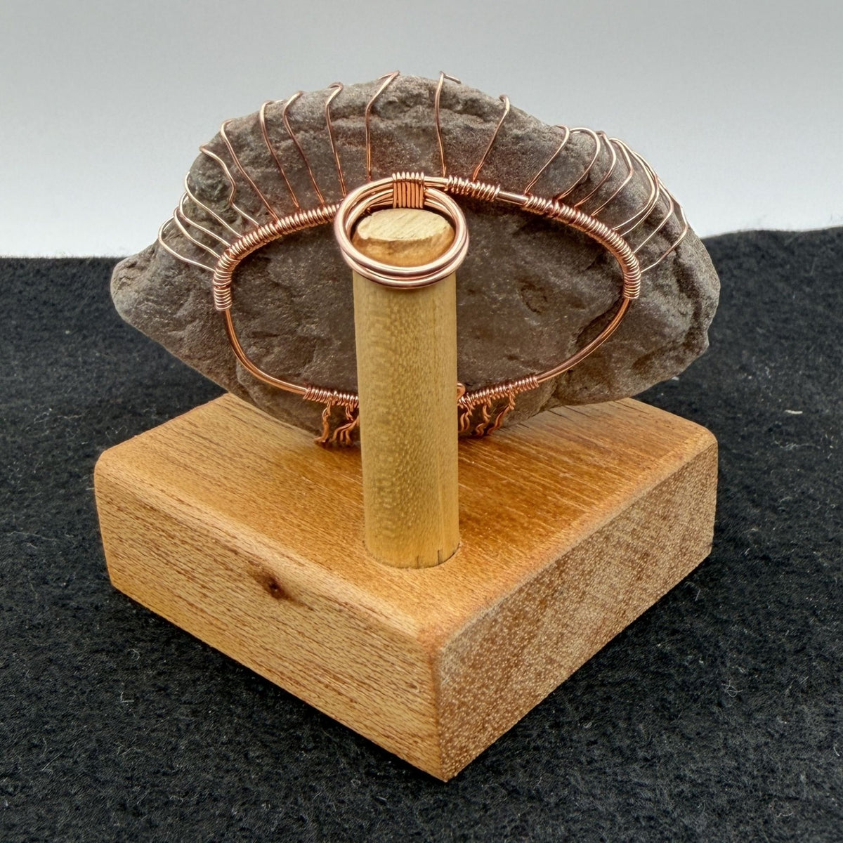 Back view of a decorative tree of life sculpture made from copper wire wrapped over a rough river stone, displayed on a rectangular light wood block against a neutral gray and black background.