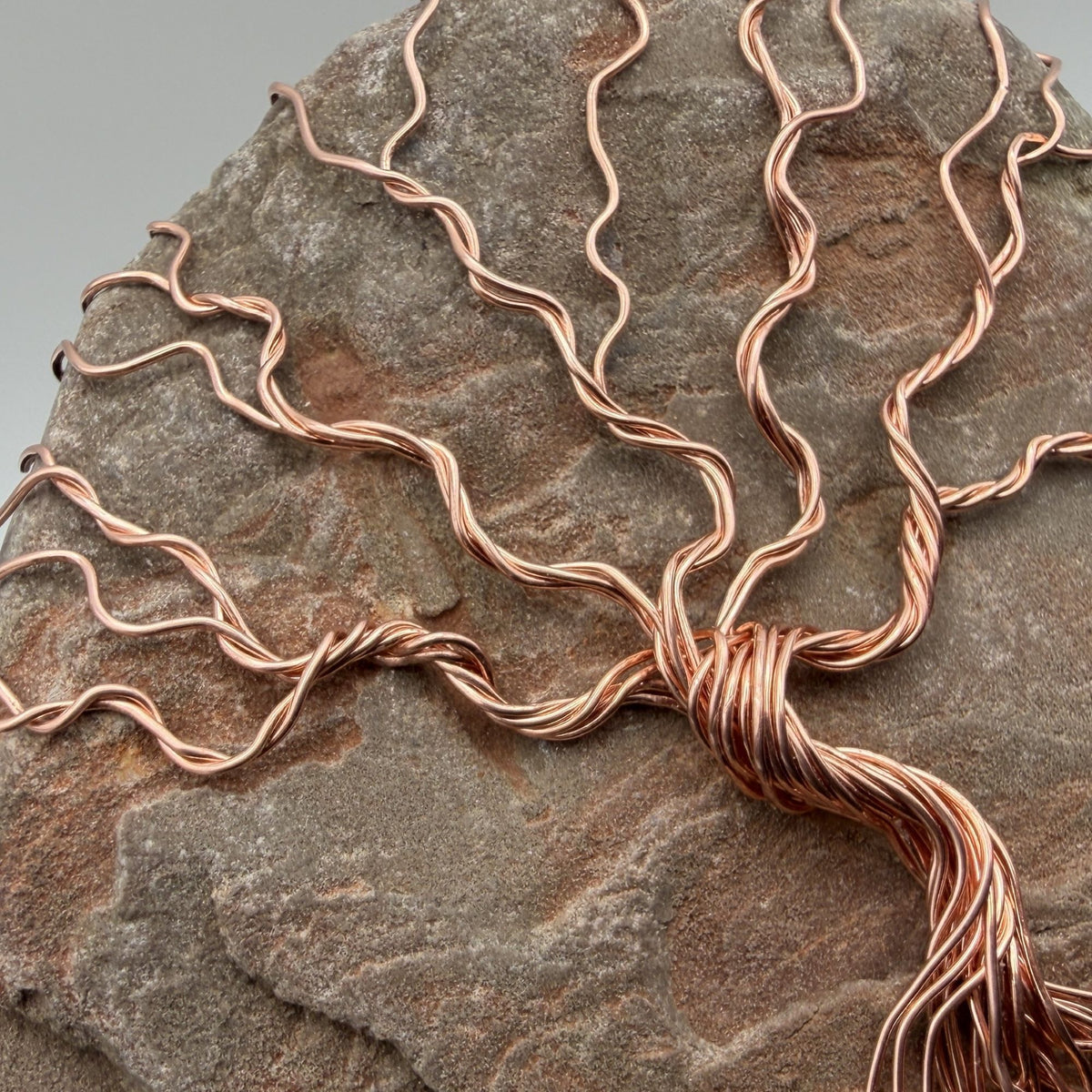 Detail view of a decorative tree of life sculpture made from copper wire wrapped over a rough river stone, displayed on a rectangular light wood block against a neutral gray and black background.