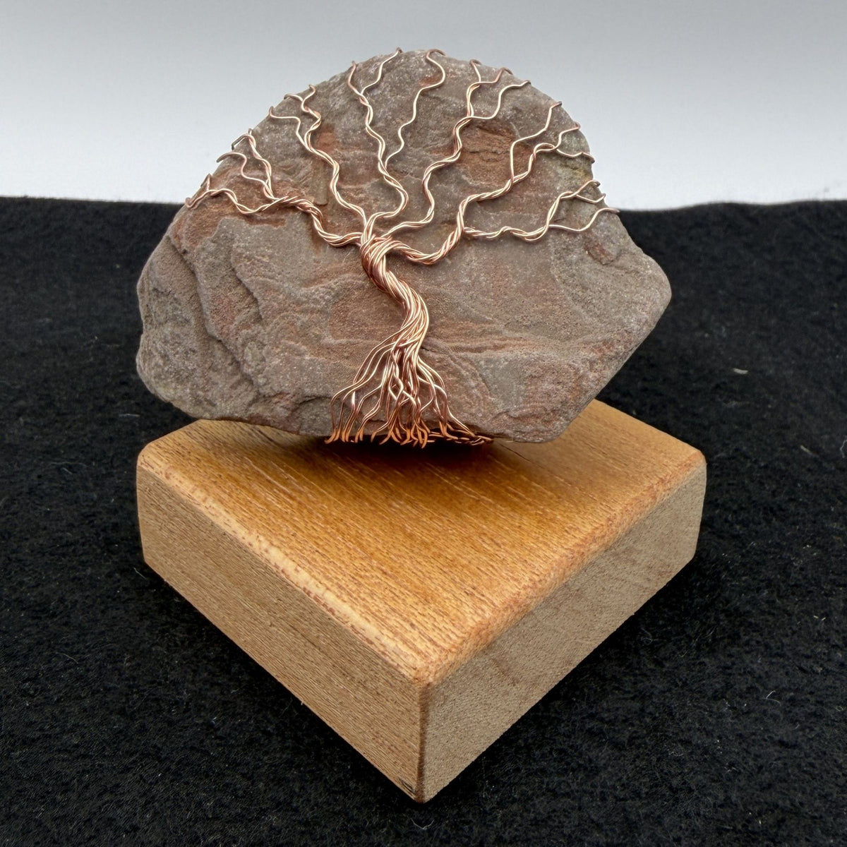 Front view of a decorative tree of life sculpture made from copper wire wrapped over a rough river stone, displayed on a rectangular light wood block against a neutral gray and black background.