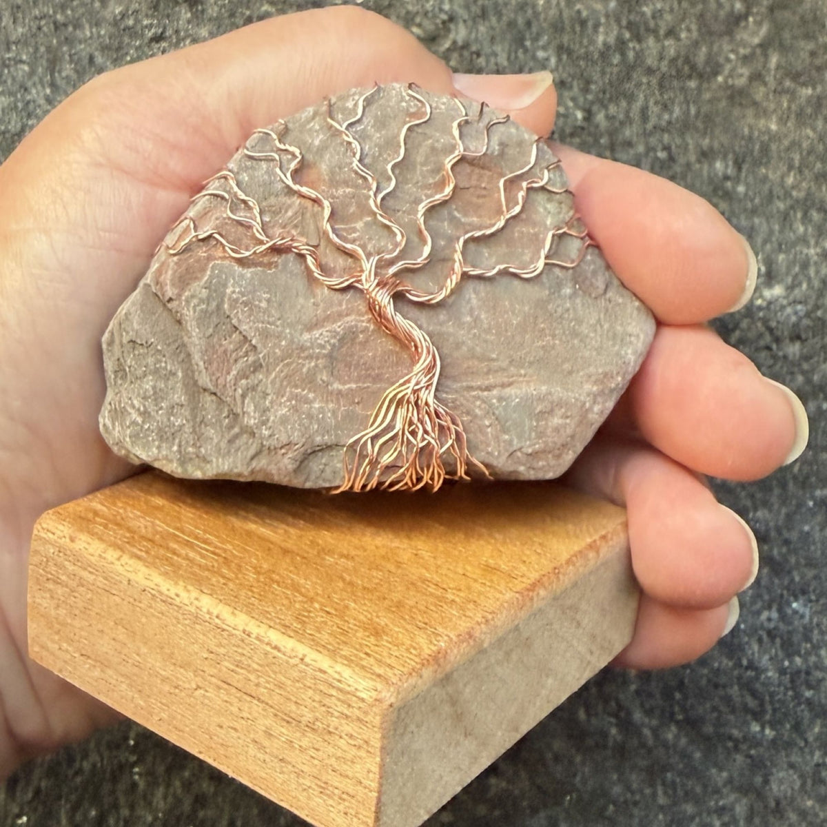 Front view of a decorative tree of life sculpture made from copper wire wrapped over a rough river stone, displayed on a rectangular light wood block and held in the artist's hand.