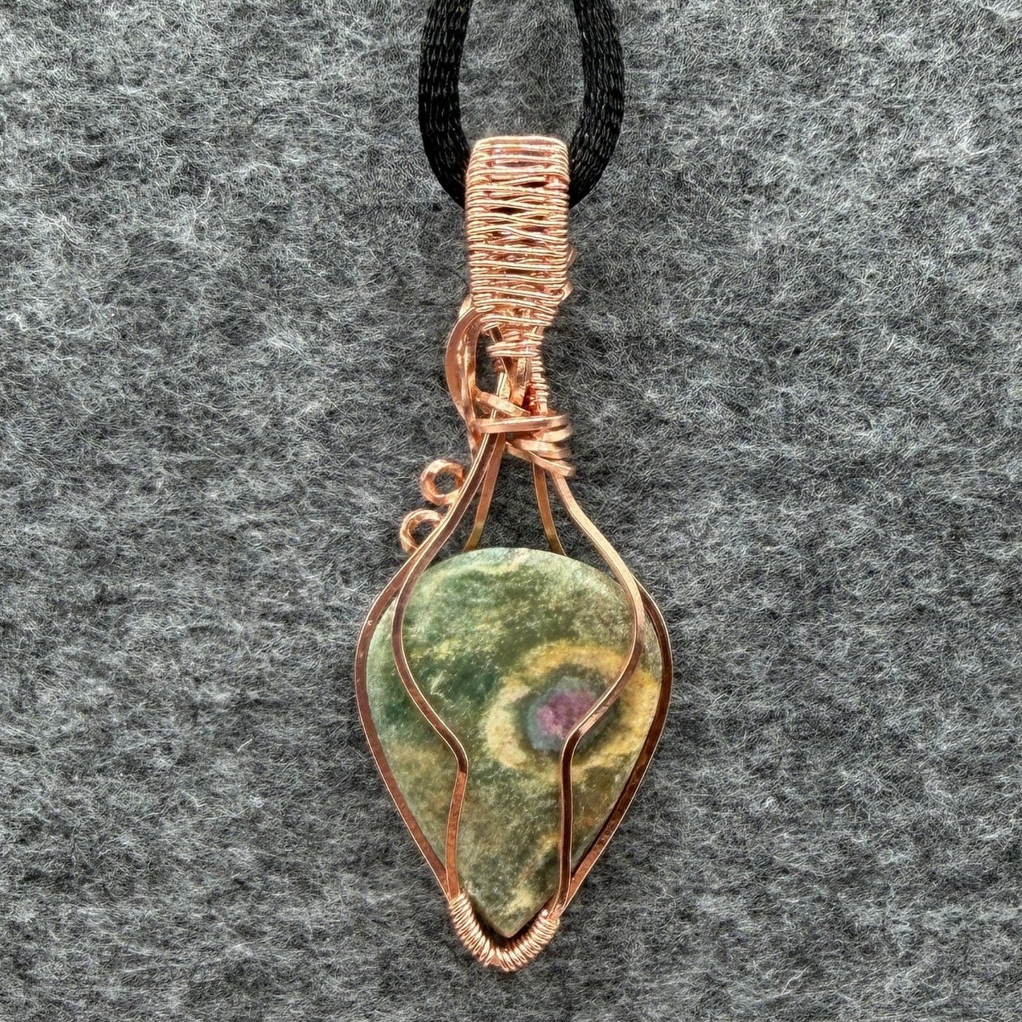 Back view of a handmade teardrop-shaped pendant necklace featuring a green ruby fuchsite stone wrapped in copper wire, suspended from a black cord, set against a textured gray background.
