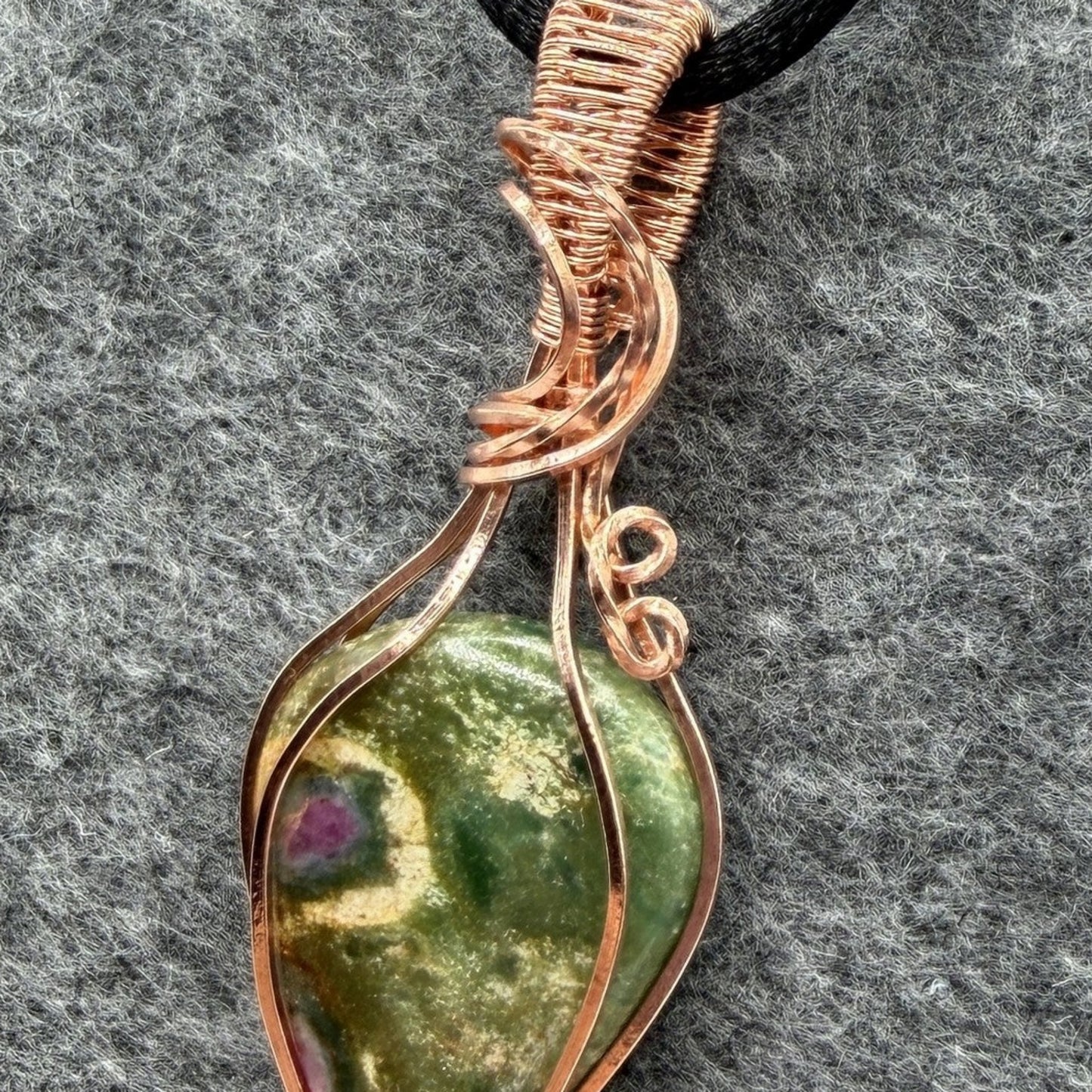 Detail view of a handmade teardrop-shaped pendant necklace featuring a green ruby fuchsite stone wrapped in copper wire, suspended from a black cord, set against a textured gray background.