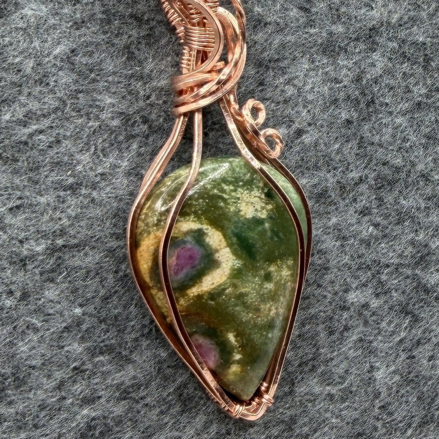 Detail view of a handmade teardrop-shaped pendant necklace featuring a green ruby fuchsite stone wrapped in copper wire, suspended from a black cord, set against a textured gray background.