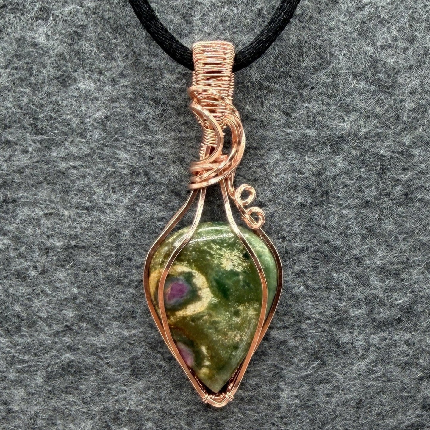 Front view of a handmade teardrop-shaped pendant necklace featuring a green ruby fuchsite stone wrapped in copper wire, suspended from a black cord, set against a textured gray background.