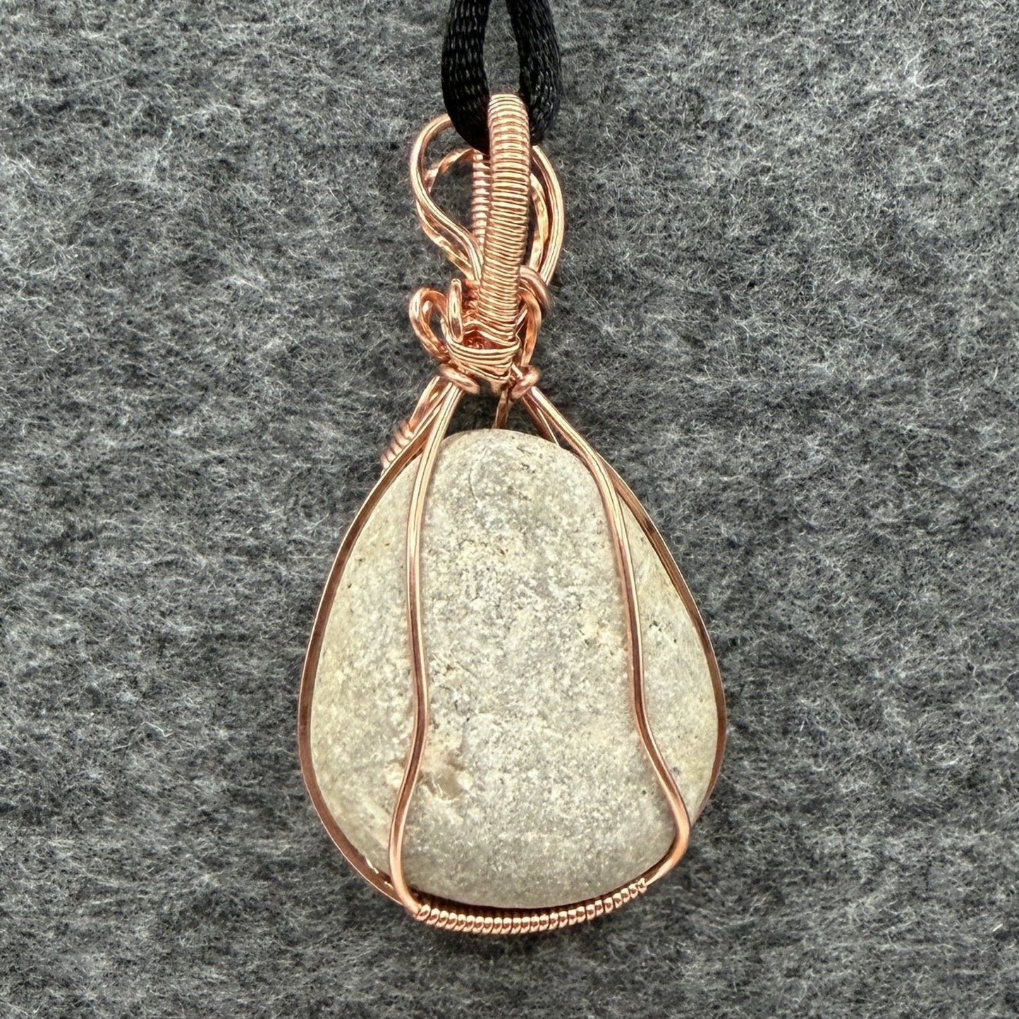 Back view of a teardrop-shaped stone pendant wrapped in copper wire, hanging from a black cord against a textured gray felt background.