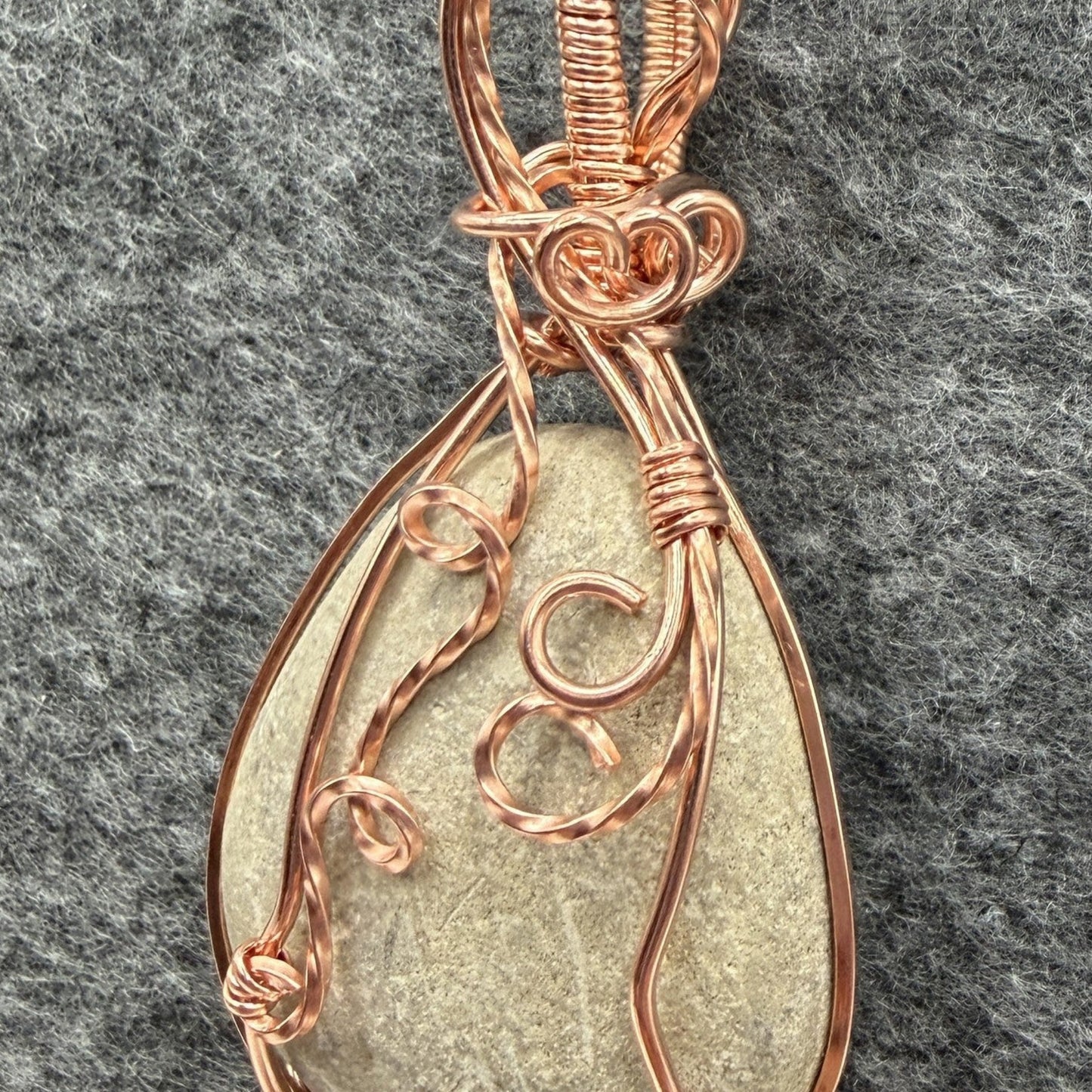 Detail view of a teardrop-shaped stone pendant wrapped in copper wire, hanging from a black cord against a textured gray felt background.