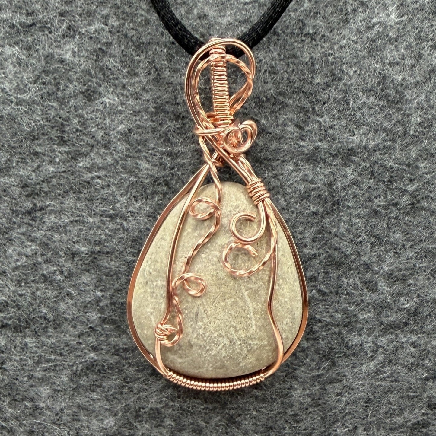 Front view of a teardrop-shaped stone pendant wrapped in copper wire, hanging from a black cord against a textured gray felt background.