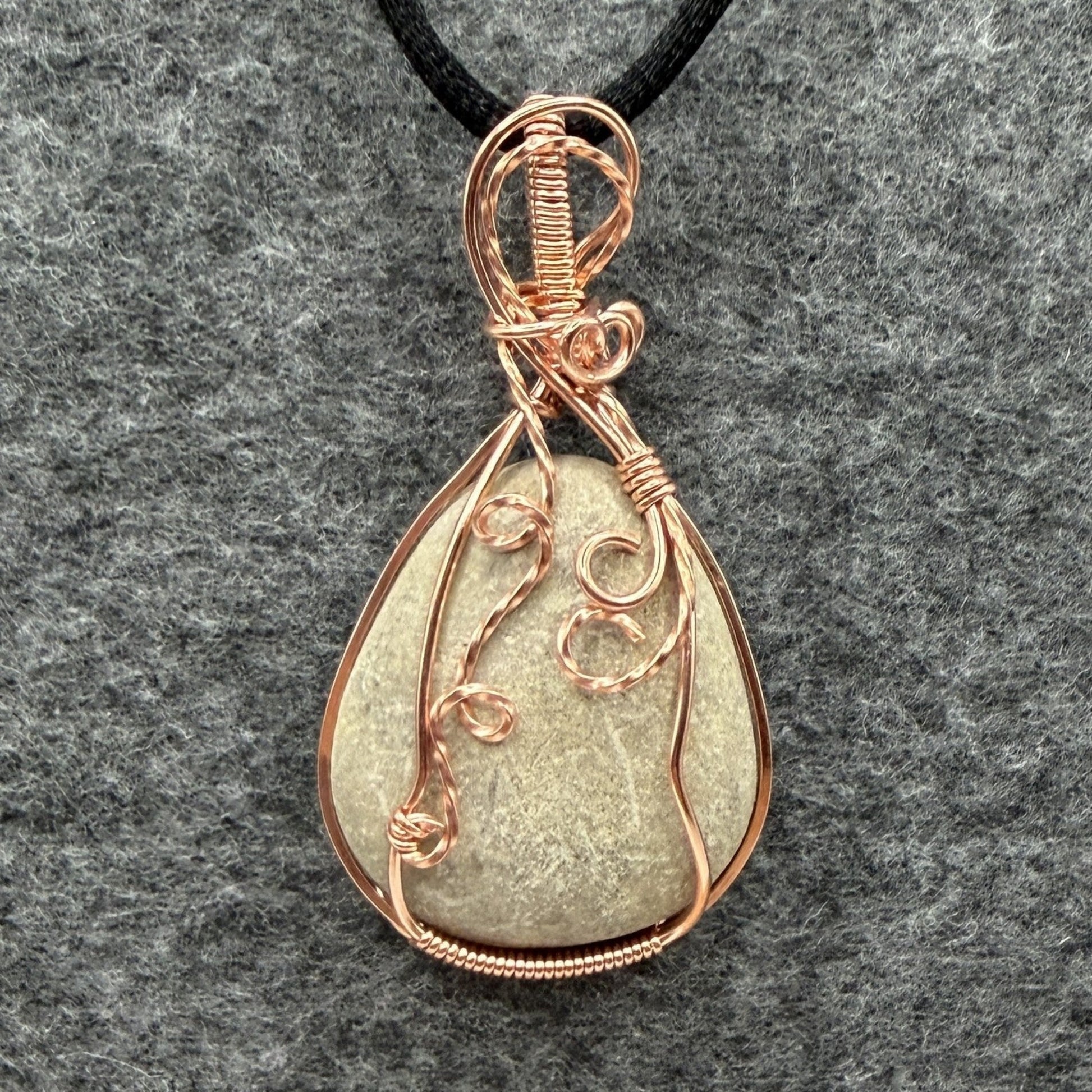 Front view of a teardrop-shaped stone pendant wrapped in copper wire, hanging from a black cord against a textured gray felt background.