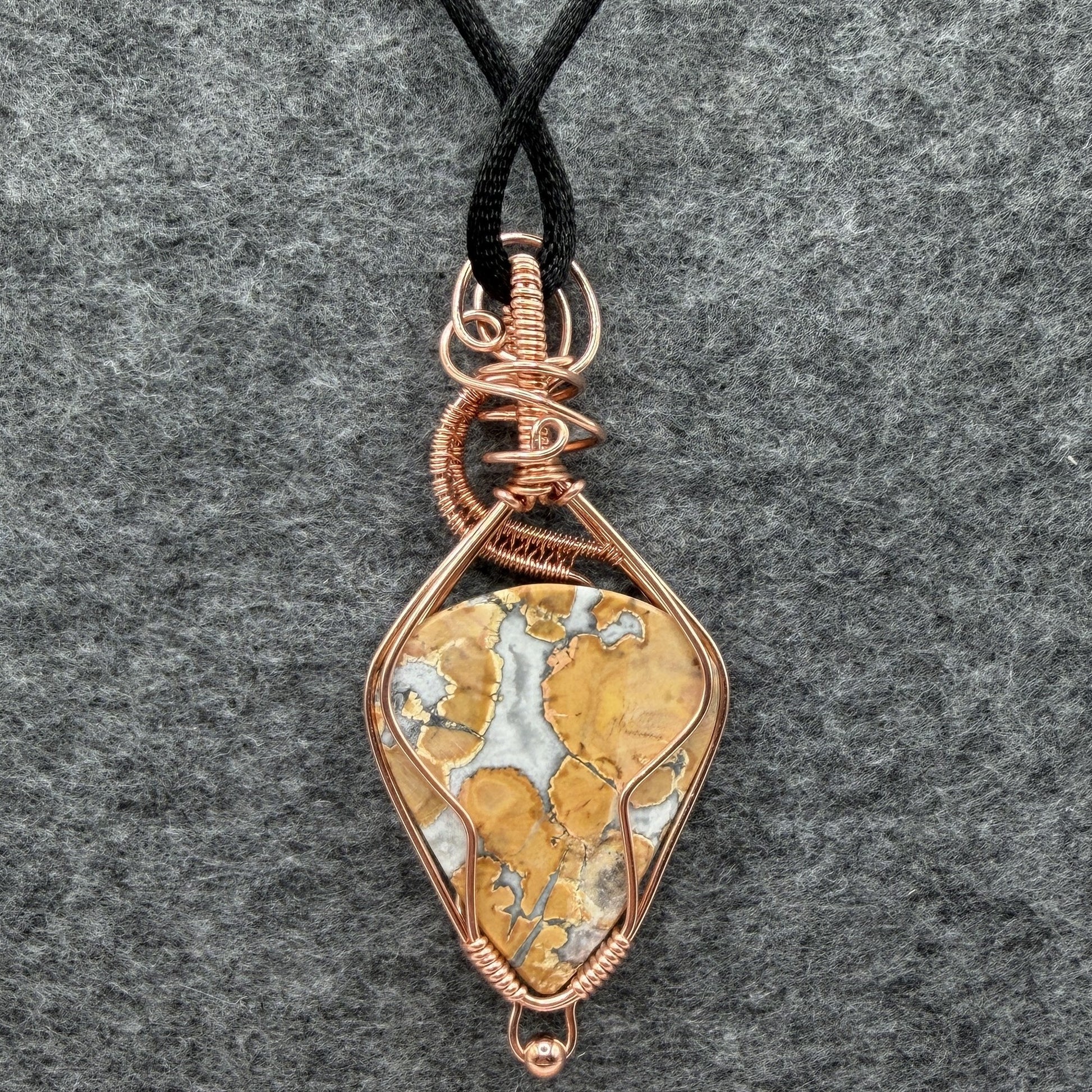 Back view of a handmade wire-wrapped pendant necklace featuring a polished yellow and white marbled maligano jasper stone, wrapped in copper wire with decorative swirls, hanging from a black cord against a textured gray background.