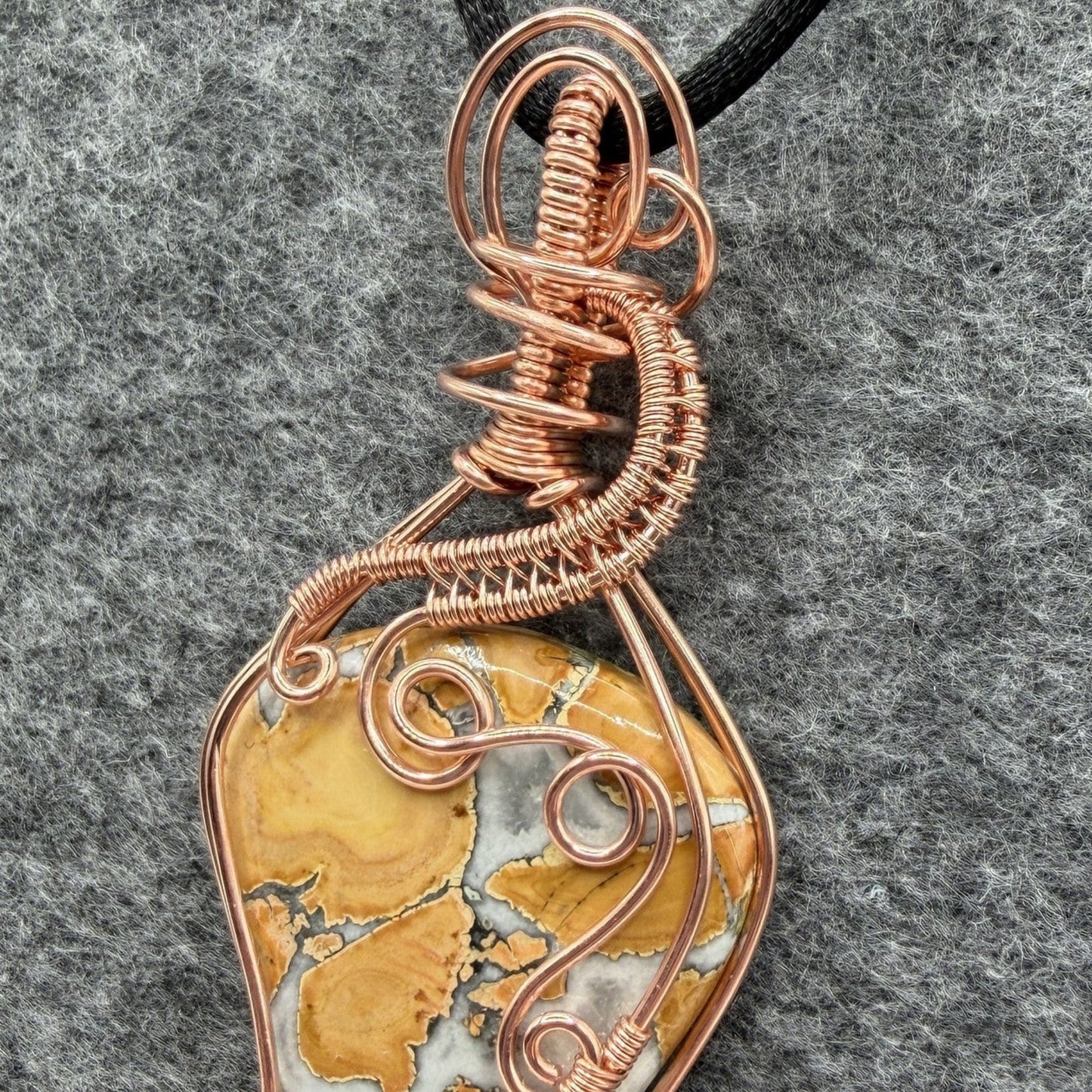 Detail view of a handmade wire-wrapped pendant necklace featuring a polished yellow and white marbled maligano jasper stone, wrapped in copper wire with decorative swirls, hanging from a black cord against a textured gray background.