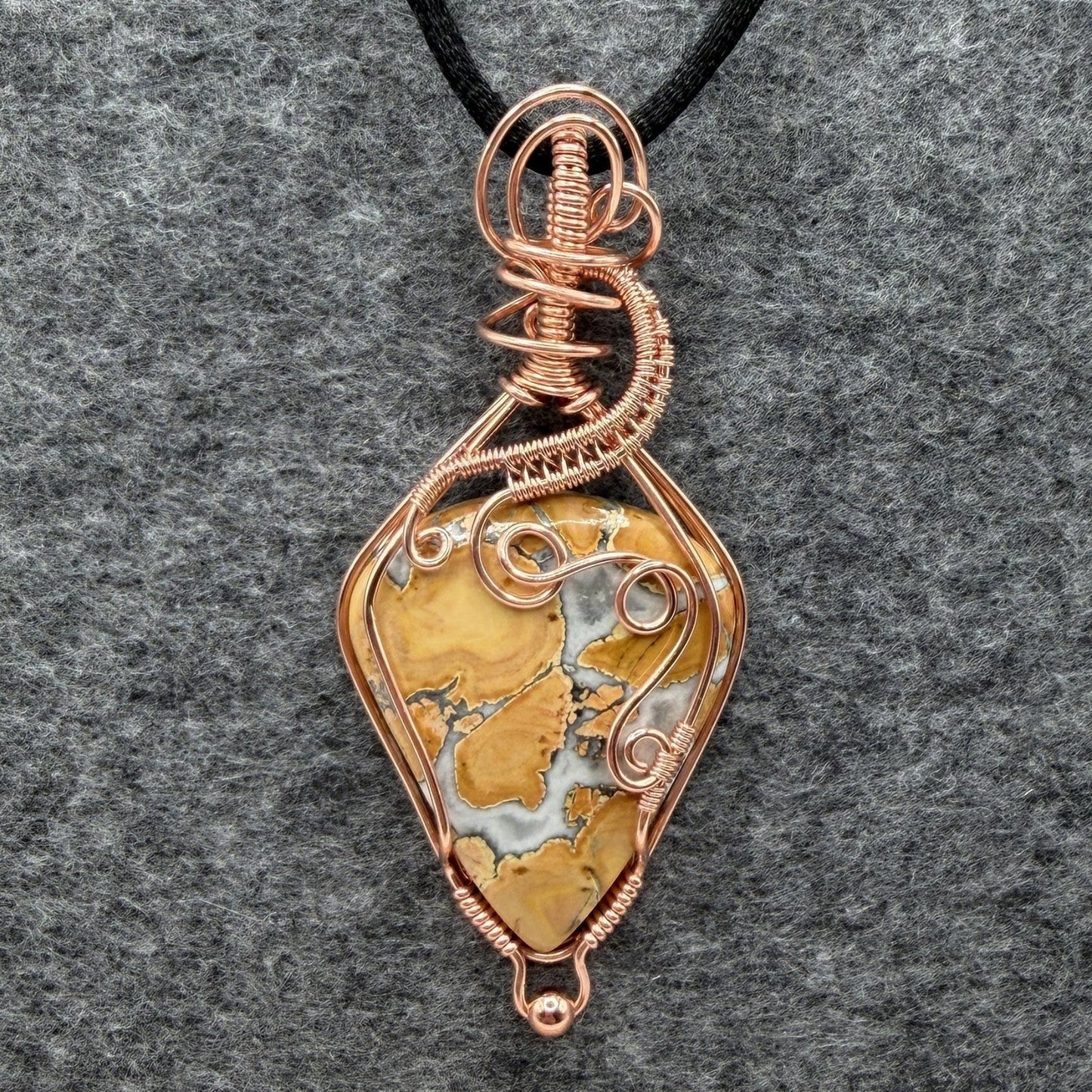 Front view of a handmade wire-wrapped pendant necklace featuring a polished yellow and white marbled maligano jasper stone, wrapped in copper wire with decorative swirls, hanging from a black cord against a textured gray background.