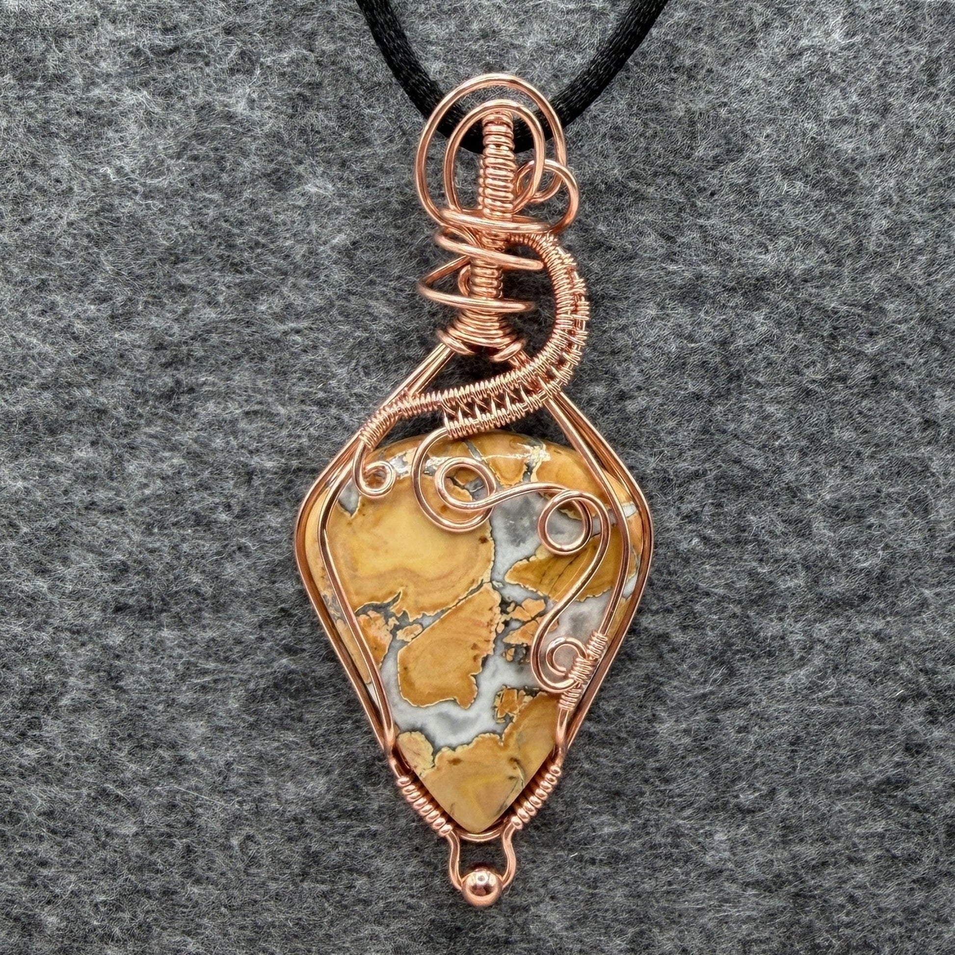 Front view of a handmade wire-wrapped pendant necklace featuring a polished yellow and white marbled maligano jasper stone, wrapped in copper wire with decorative swirls, hanging from a black cord against a textured gray background.