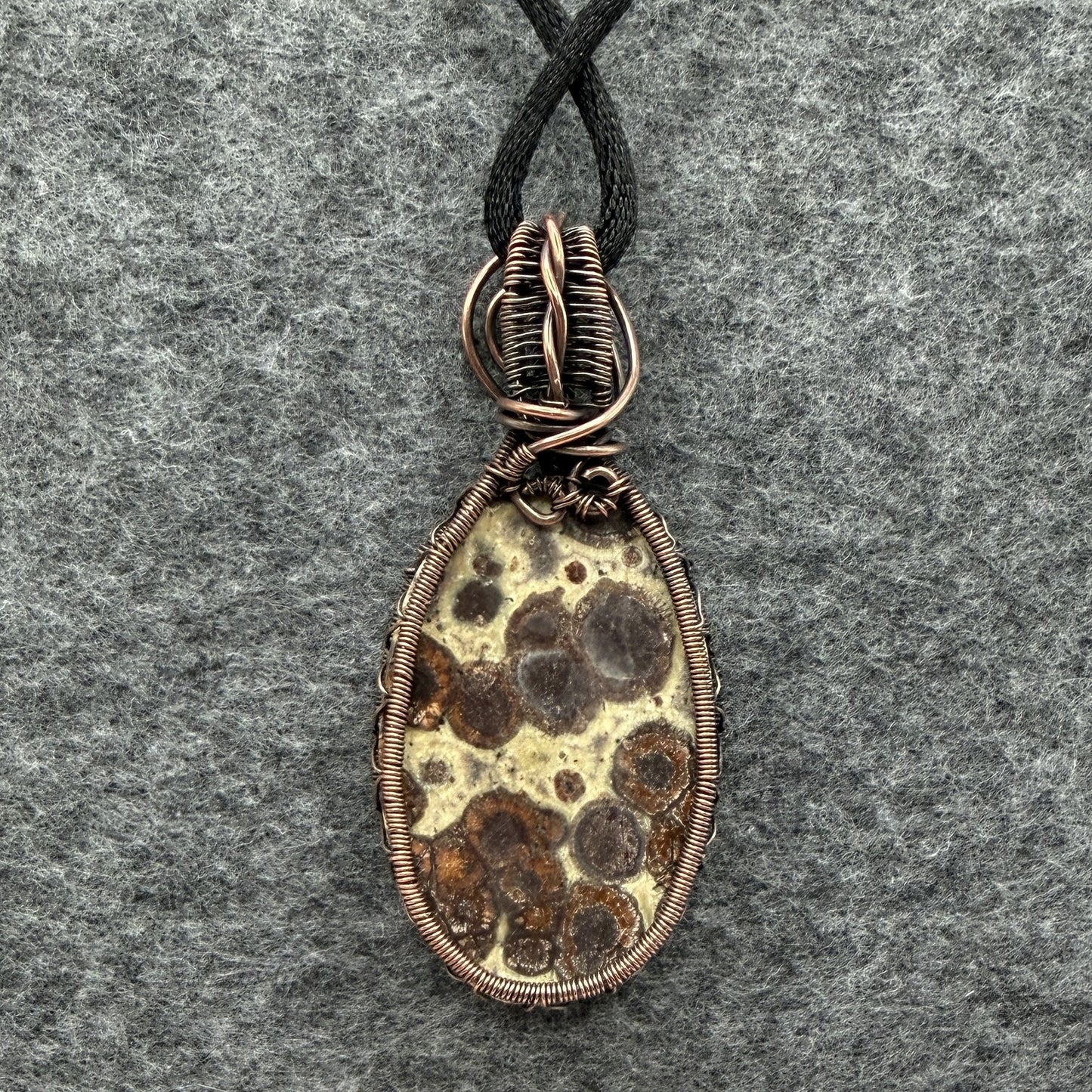 Back view of a handmade oval pendant necklace featuring a natural brown and cream asteroid jasper stone, wrapped in antiqued copper wire with decorative swirls, hanging from a black cord on a textured gray background.