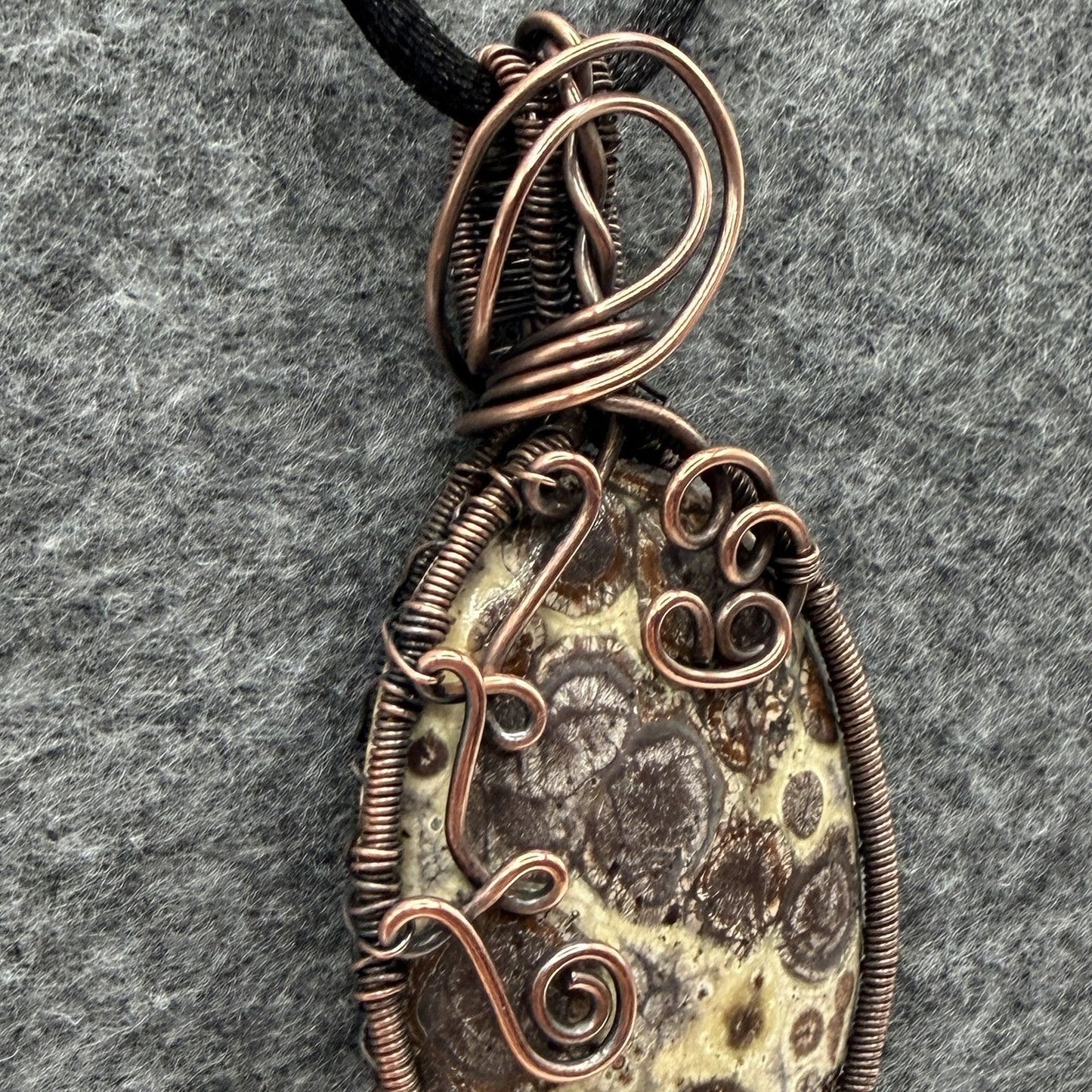 Detail view of a handmade oval pendant necklace featuring a natural brown and cream asteroid jasper stone, wrapped in antiqued copper wire with decorative swirls, hanging from a black cord on a textured gray background.
