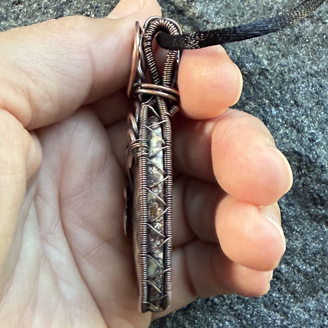 Detail view of a woven-framed handmade oval pendant necklace featuring a natural brown and cream asteroid jasper stone, wrapped in antiqued copper wire with decorative swirls, hanging from a black cord, held in the artist's hand.